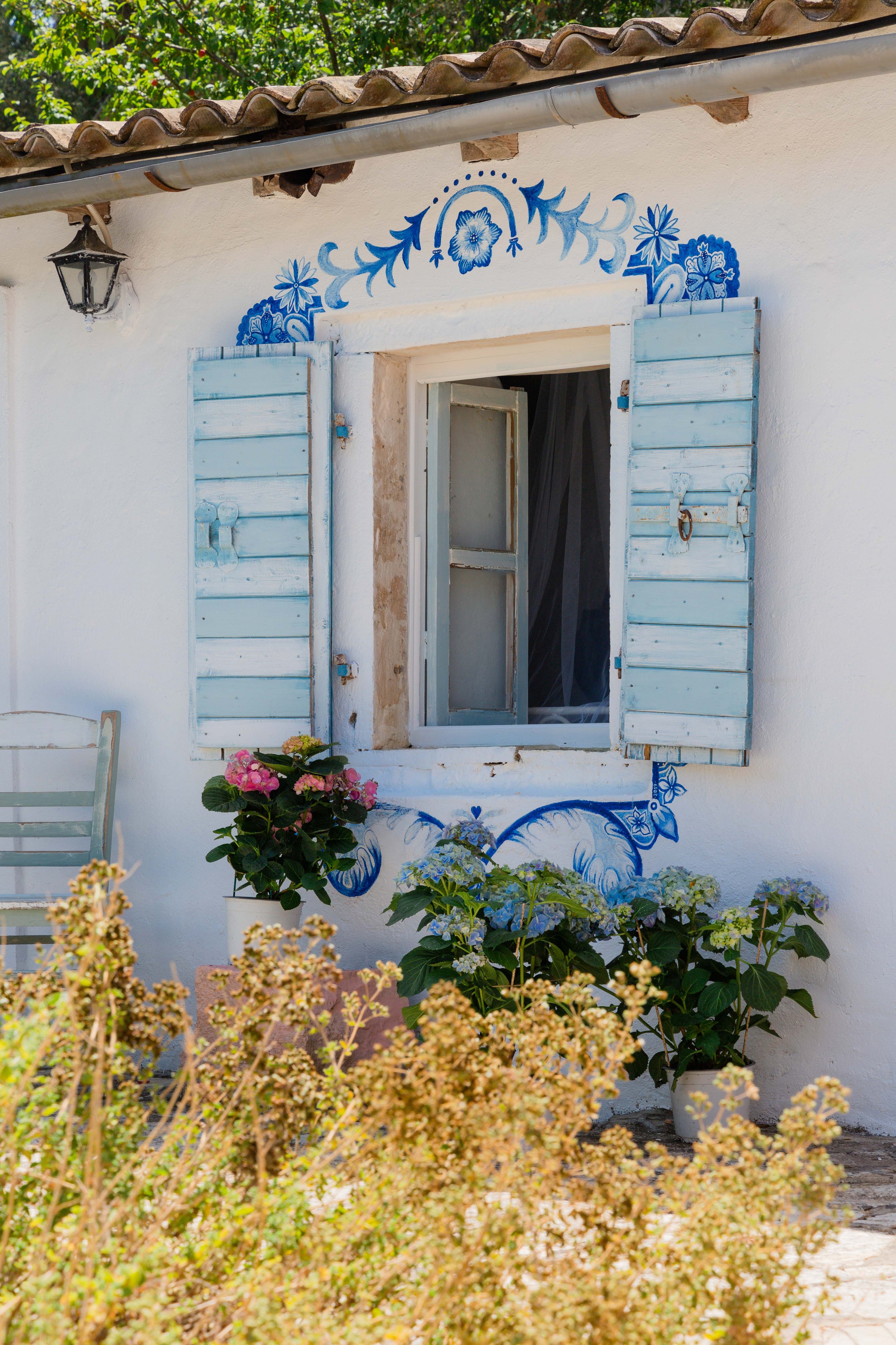 A white house wall with a blue and white painted floral design around a window with open light blue wooden shutters, a black outdoor lamp, and potted plants with pink, white, and light purple flowers in front.