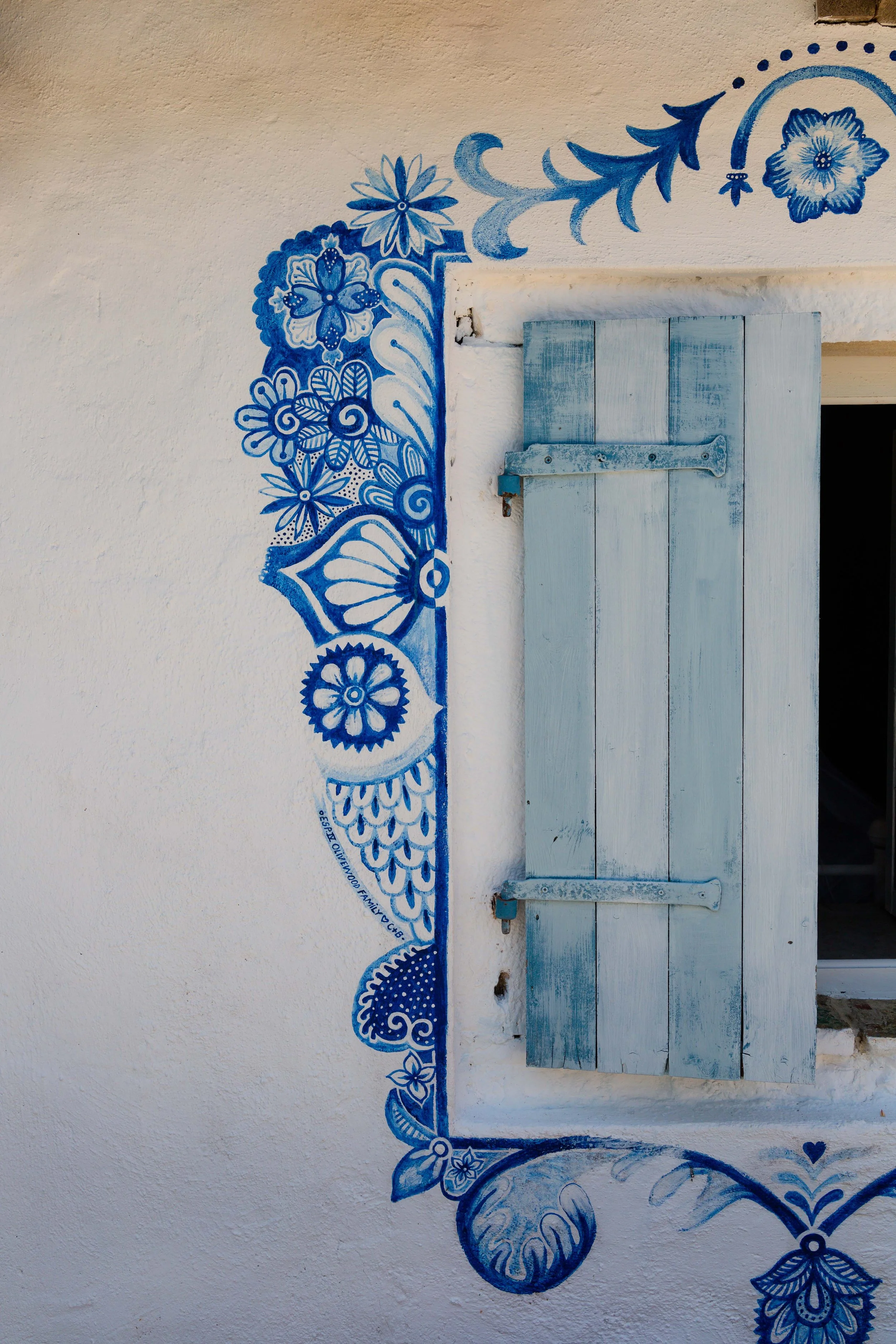 White stucco wall with blue floral and decorative mural next to a small closed blue wooden window shutter.