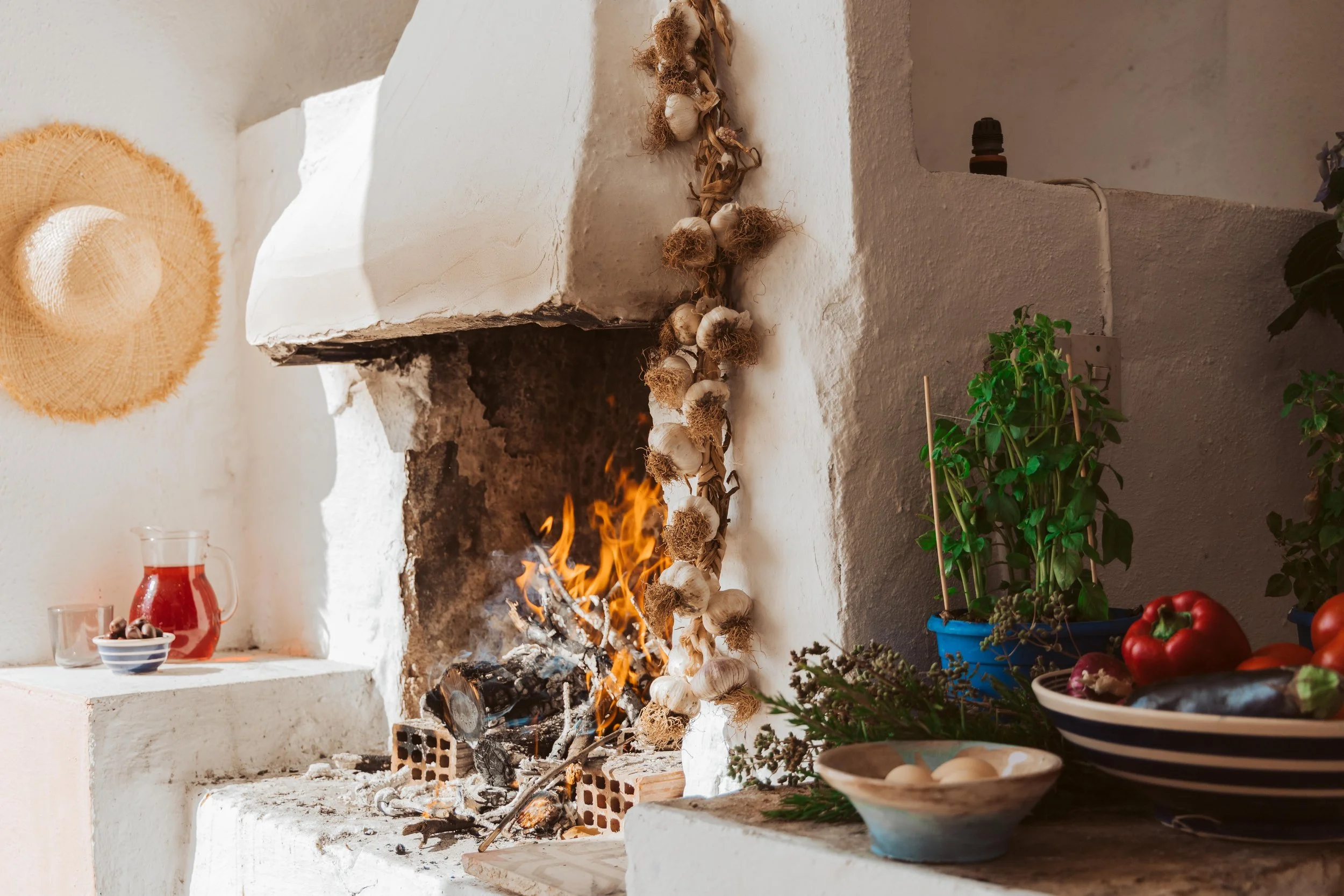 A cozy kitchen with a white plaster fireplace, a fire burning inside, decorated with dried garlic hanging on the side, and various potted herbs, vegetables, and a bowl of eggs on the kitchen counter.