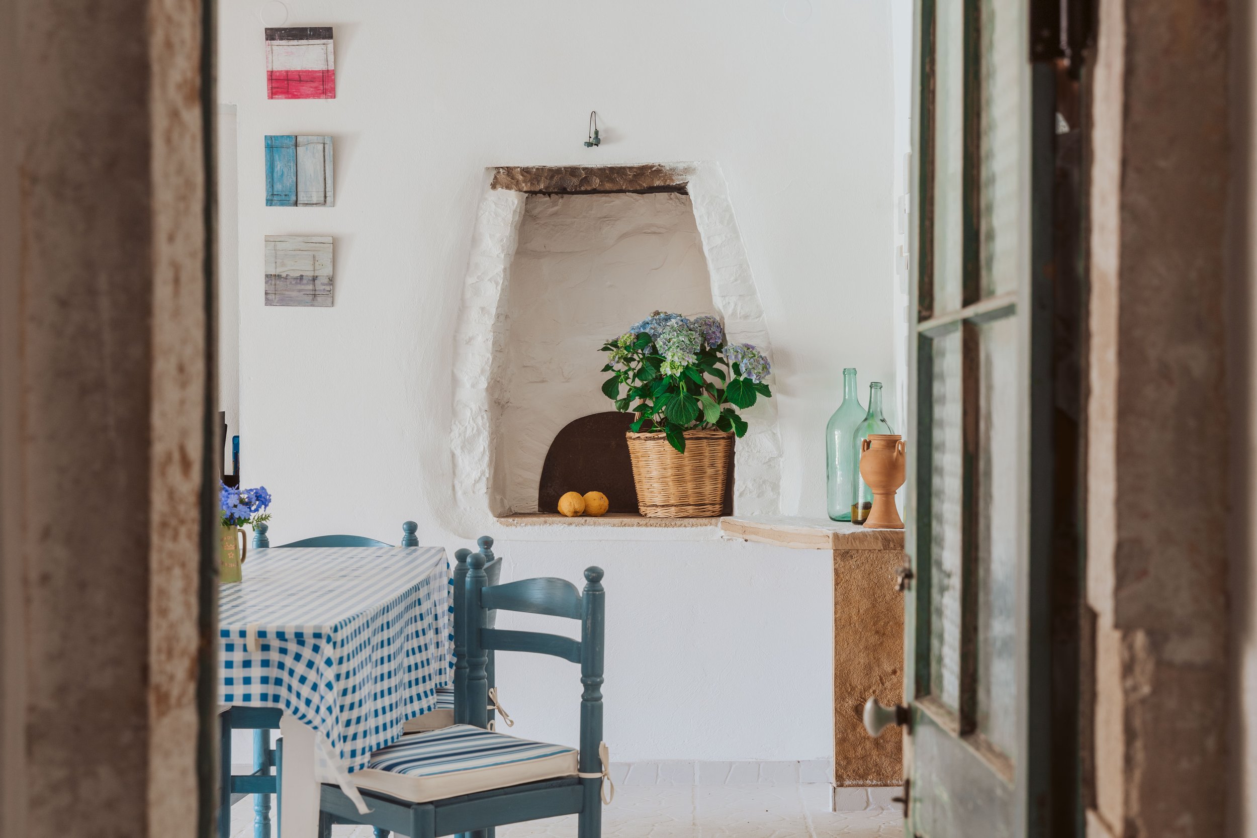 Interior view of a cozy dining area with a blue and white checkered tablecloth, blue chairs, and decorative items like a potted hydrangea, glass bottles, and ceramics, seen through an open door.