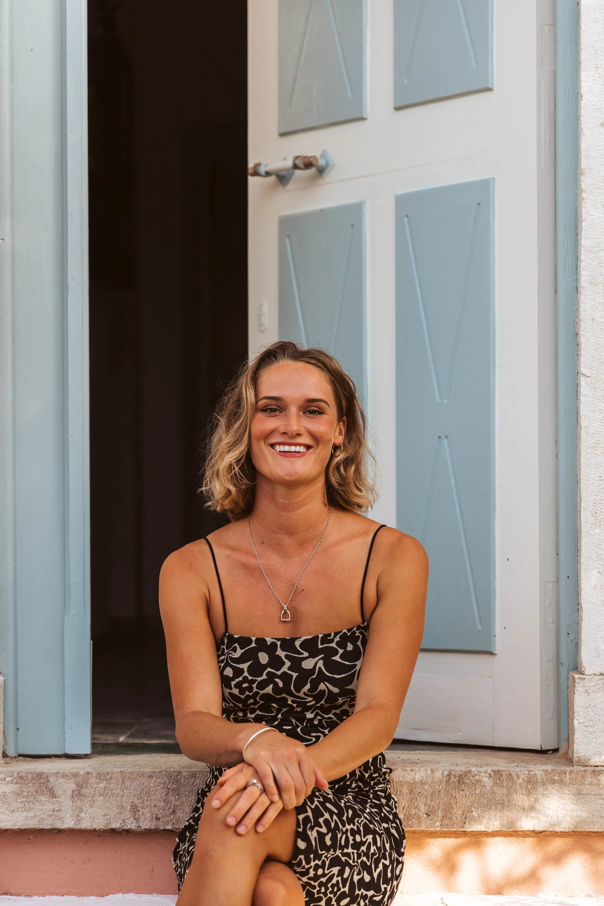A young woman with blonde hair, wearing a black and beige patterned dress, sitting on the front step of a house with light blue shutters and a white door, smiling at the camera.