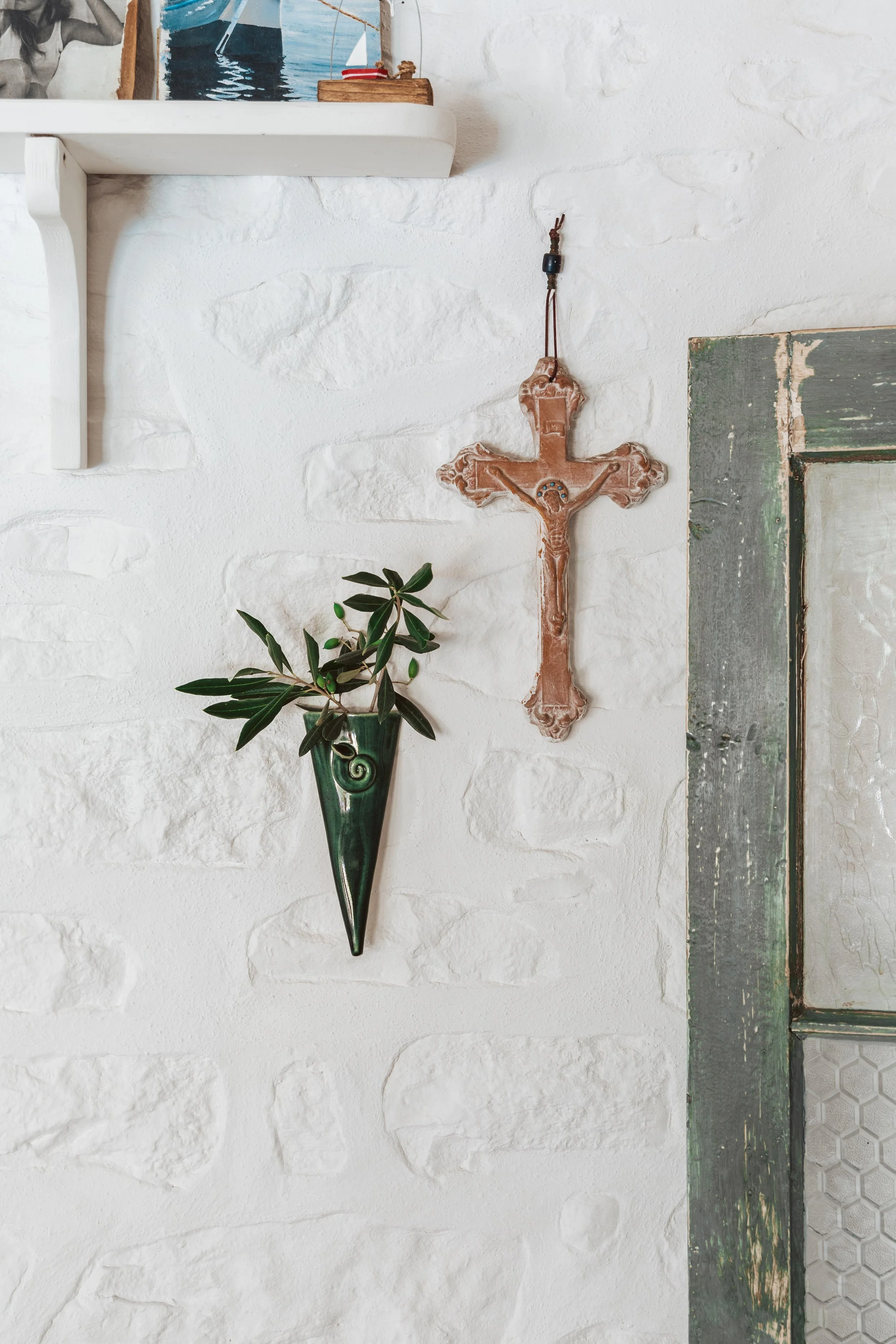 Decorative wall with a terracotta cross hanging and a green potted plant in a green wall-mounted cone-shaped holder, next to a rustic wooden door frame.