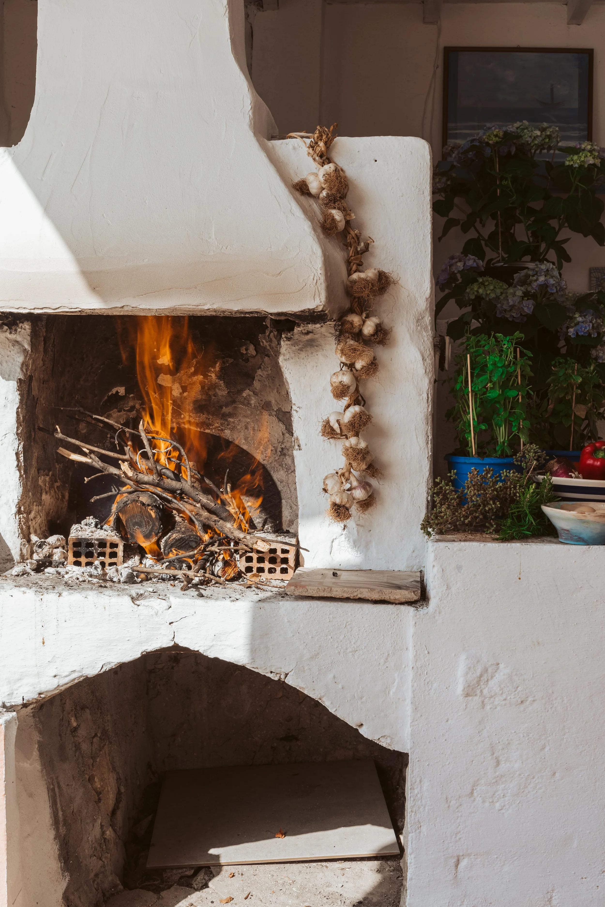 A rustic outdoor fireplace with a fire burning, decorated with a garlic braid hanging on the side, and plants and flowers nearby.