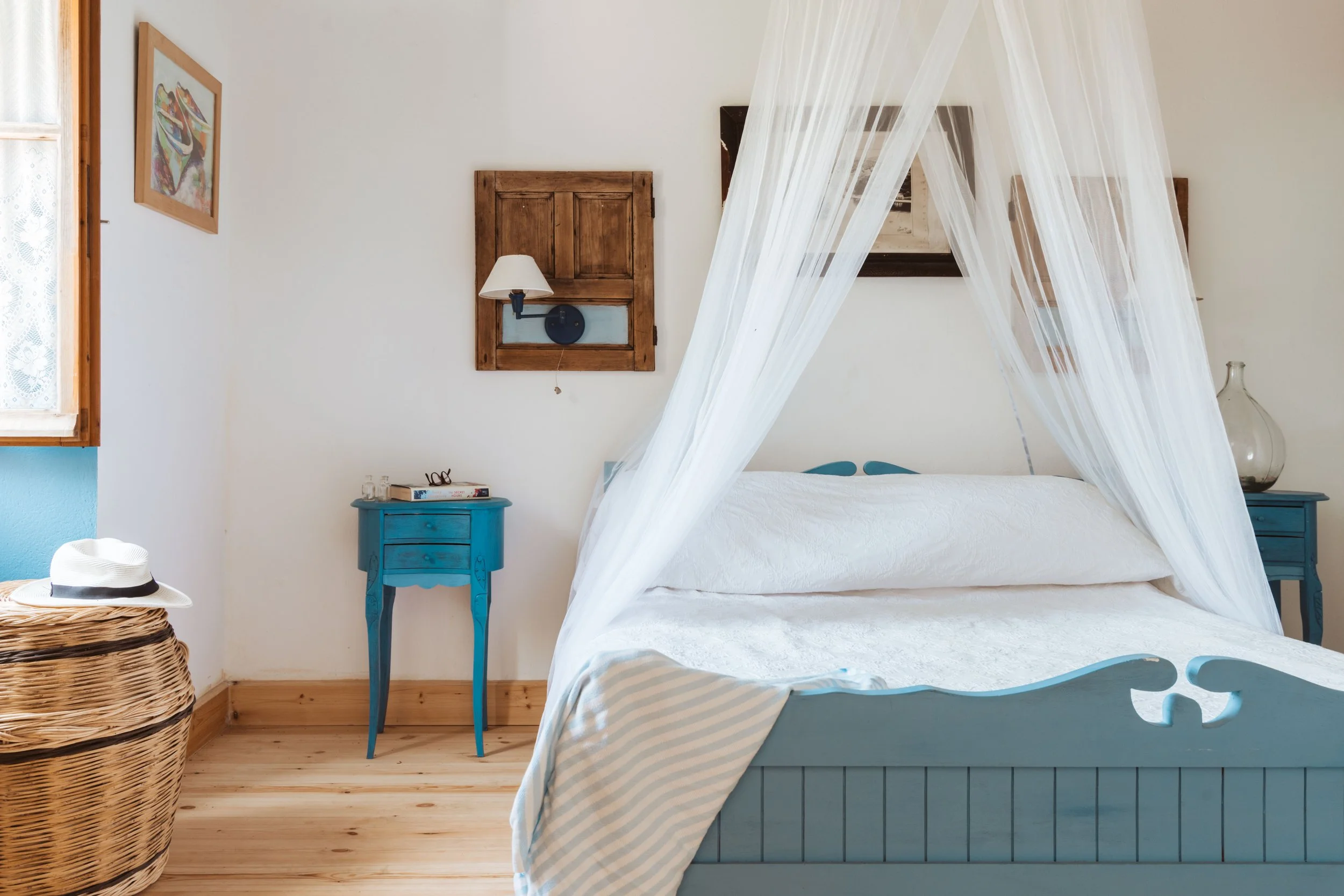 A bedroom with a blue wooden bed, white canopy, blue nightstands, wicker laundry basket and a window with lace curtains.