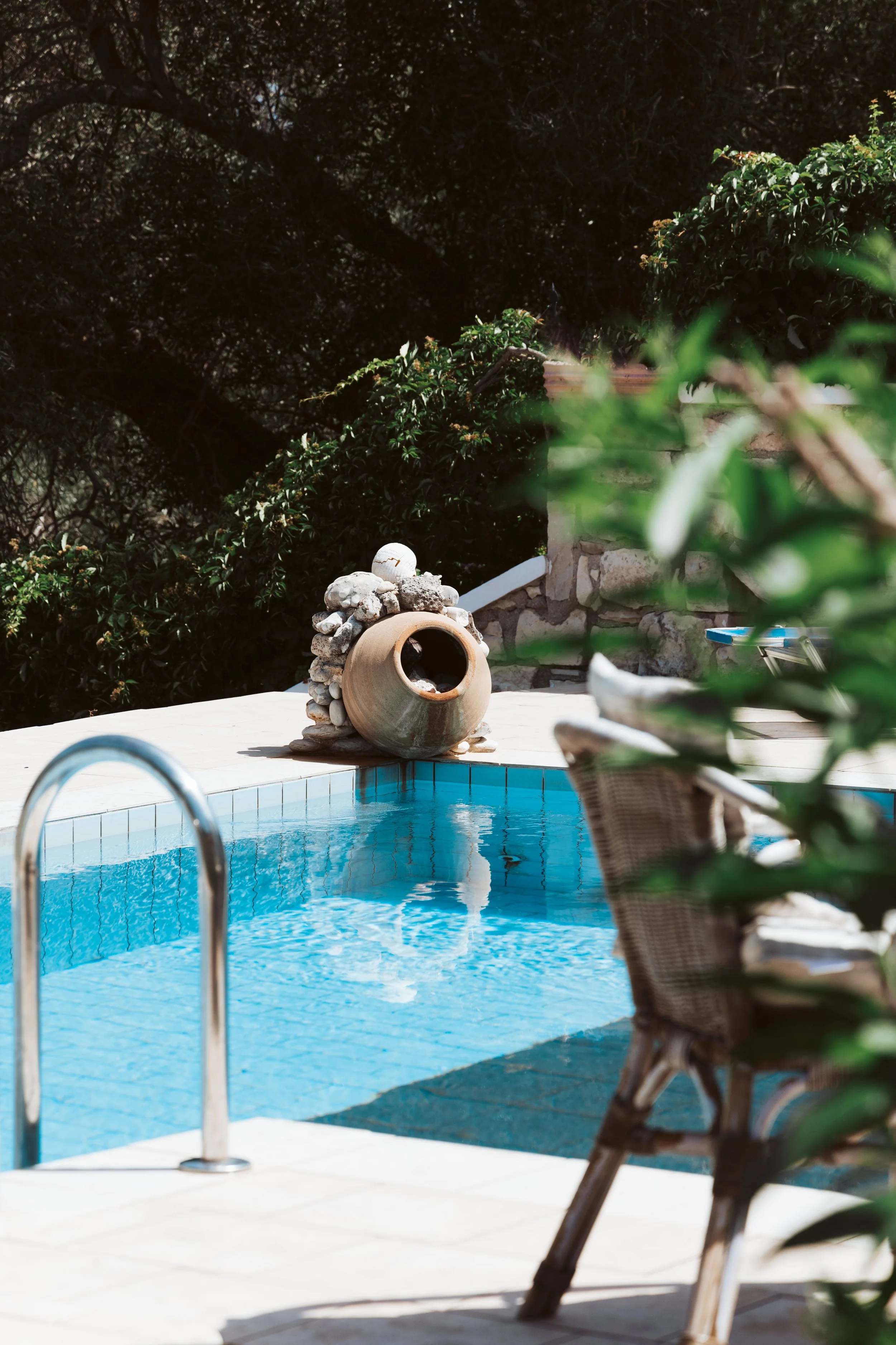 Pool with a ladder, wicker chair, and a large clay pot on the pool deck surrounded by green bushes and trees.