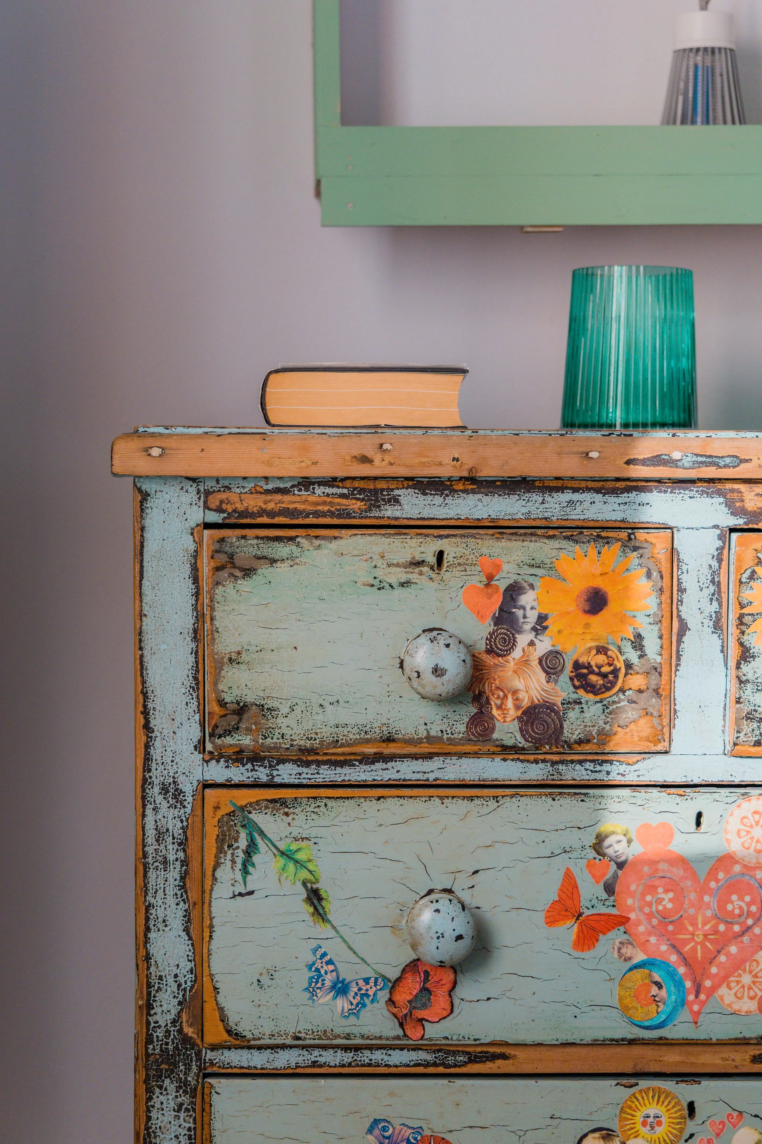 Close-up of an old, distressed light blue wooden dresser with stickers of flowers, faces, hearts, and butterflies on it. On top of the dresser, there is a closed book, a green glass lamp, and a partial view of a mirror with a green frame.