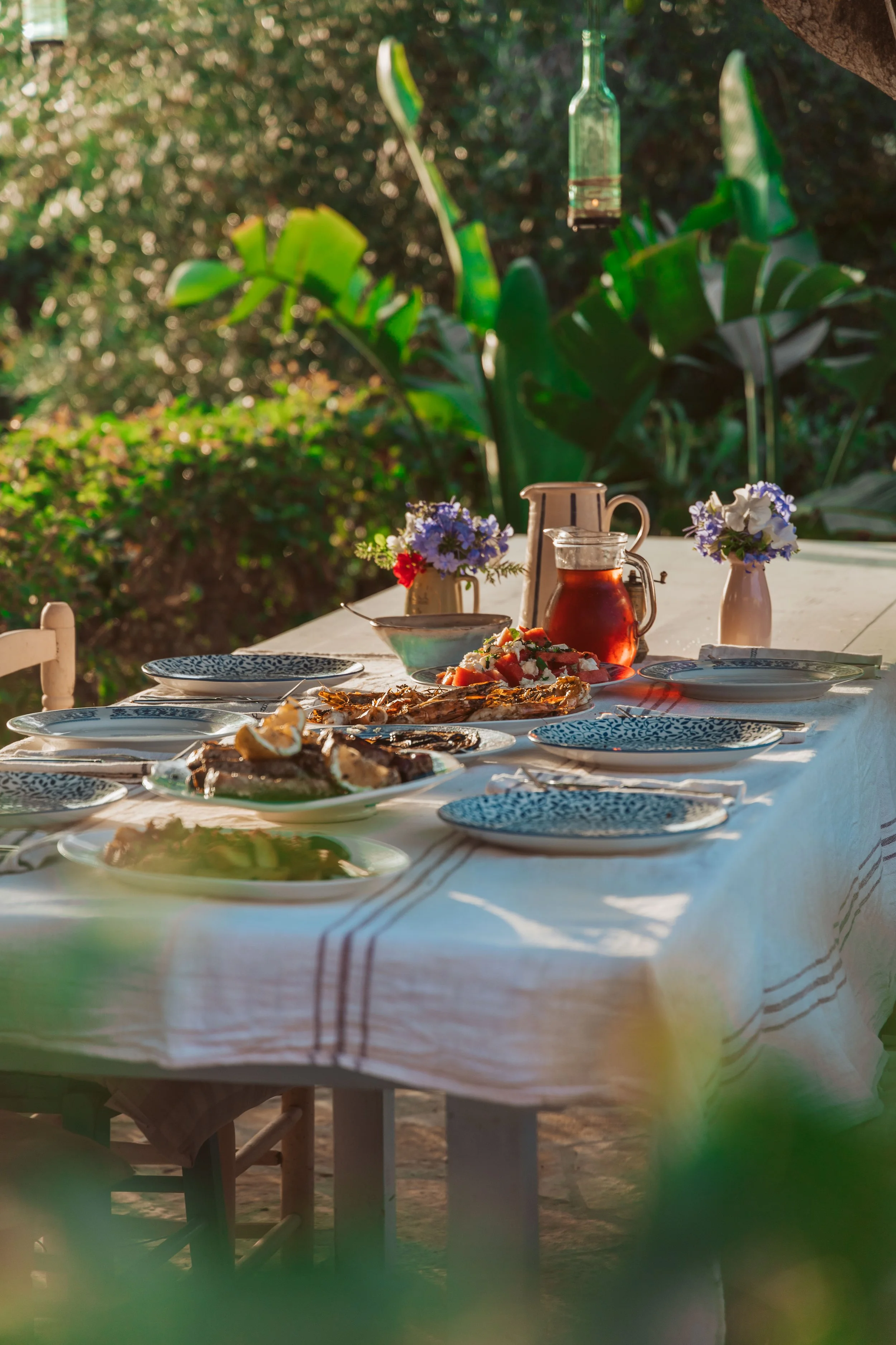 Outdoor dining table set with plates, glasses, and food, surrounded by lush greenery.
