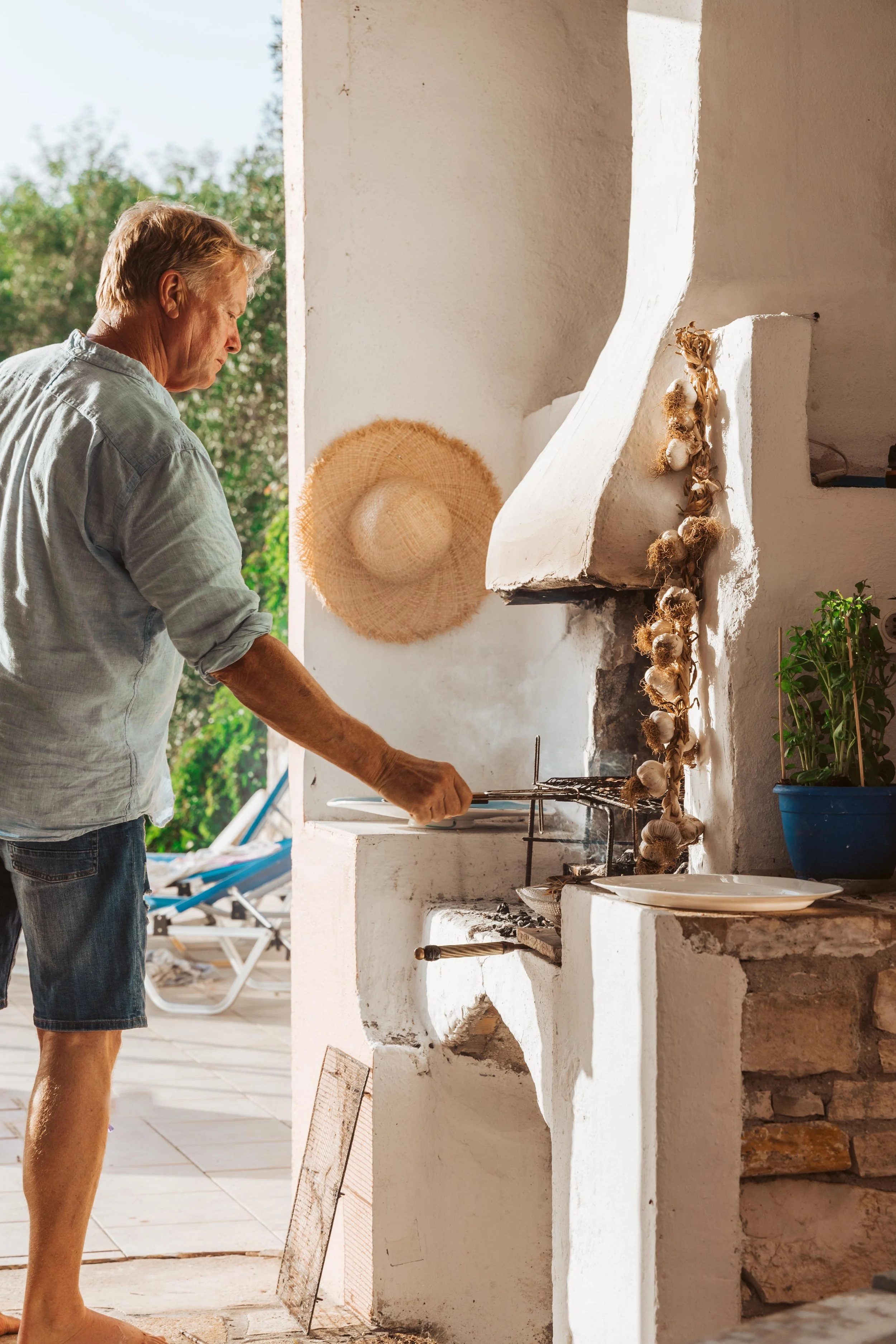 An elderly man with blonde hair is grilling food on an outdoor fireplace or grill. He is wearing a light-colored shirt and denim shorts. The scene is sunlit with greenery in the background, and there is a straw hat hanging on the wall, along with a b