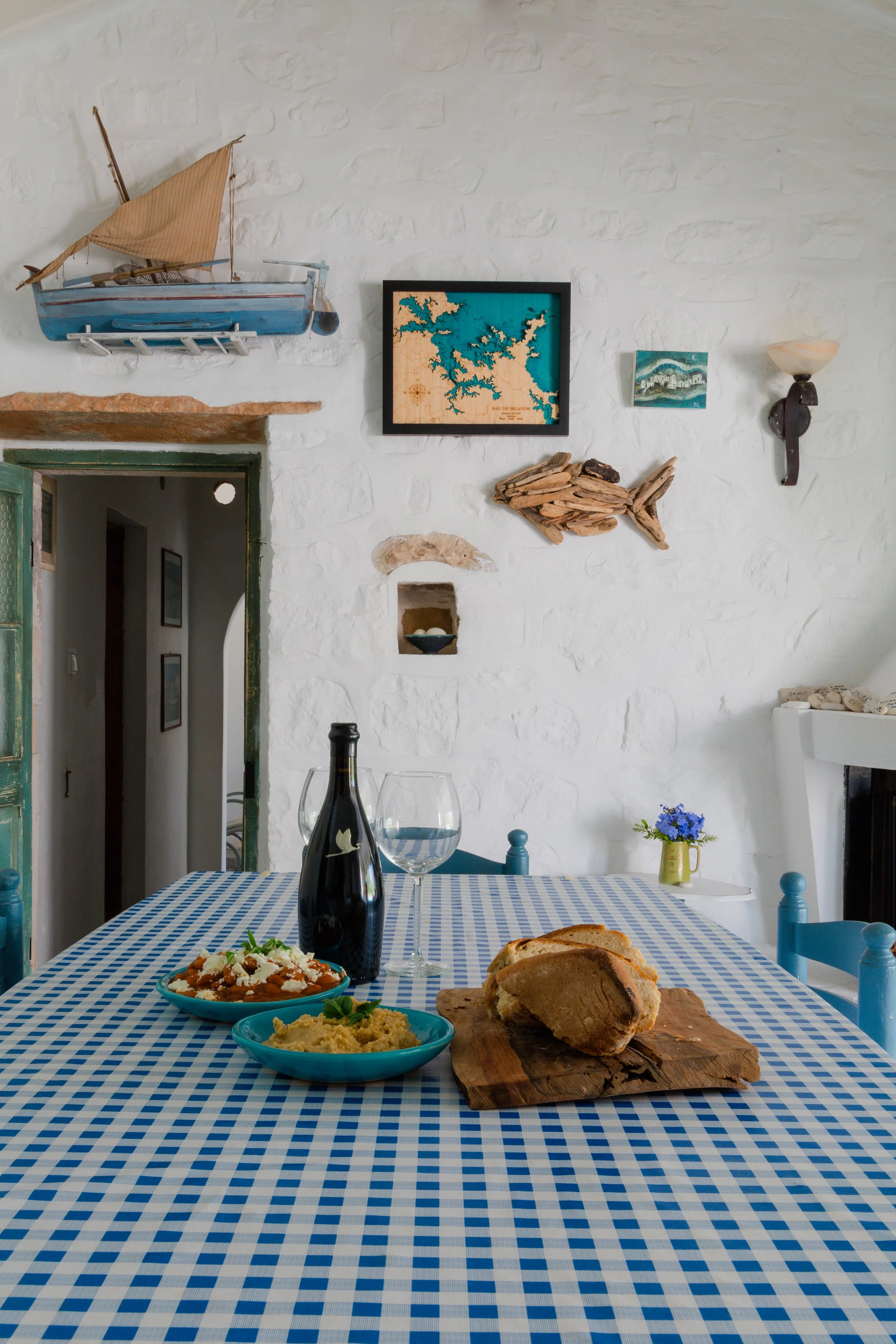 A dining table with a blue checkered tablecloth set with two bowls of food, a bottle, and two wine glasses in a room with white brick walls decorated with nautical decorations and a small vase with blue flowers.