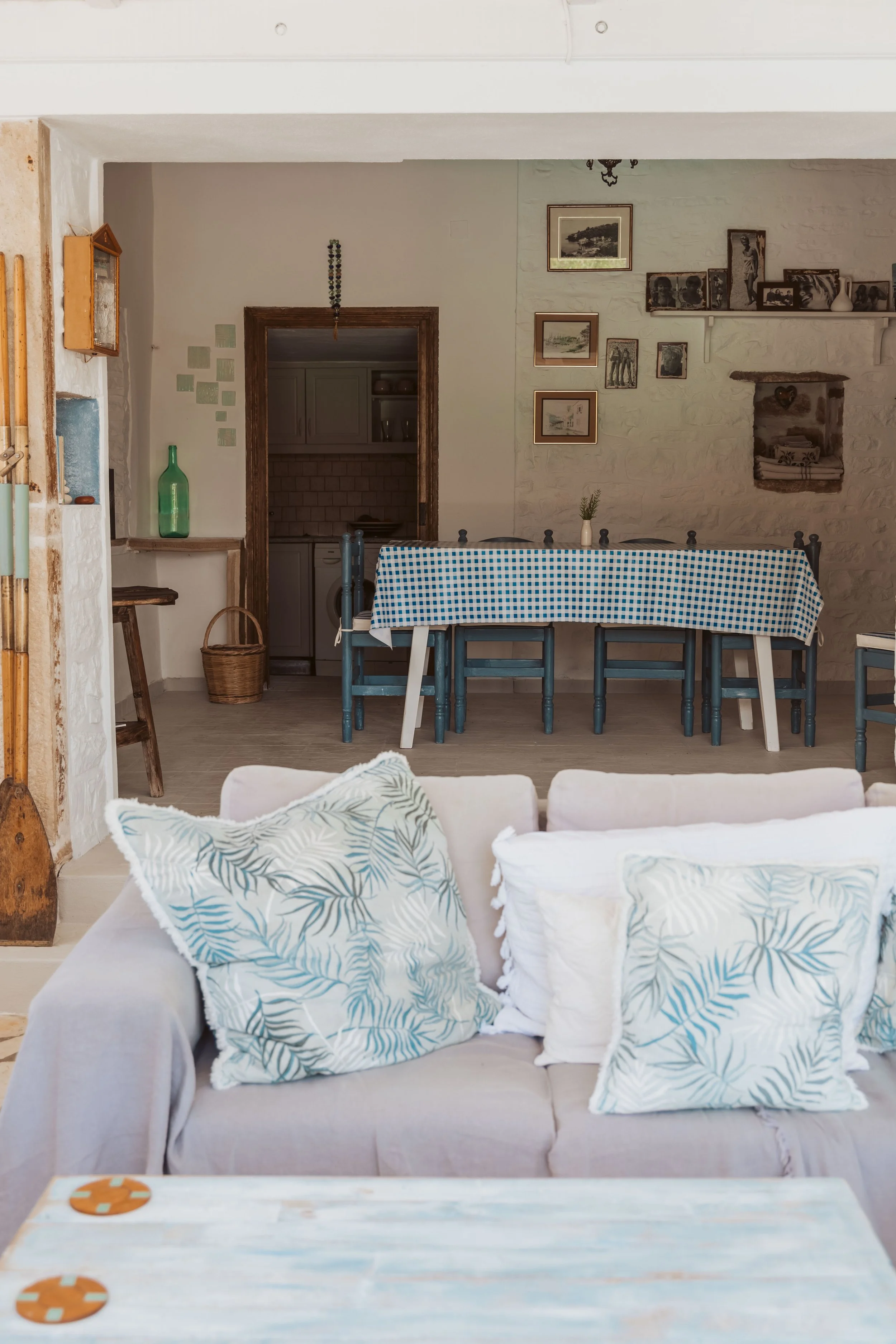 Living room with a beige sofa, decorated with tropical-patterned pillows, in the foreground. A dining table with a blue checkered tablecloth and several blue chairs is in the background, with a small white vase and greenery on it. The room features w