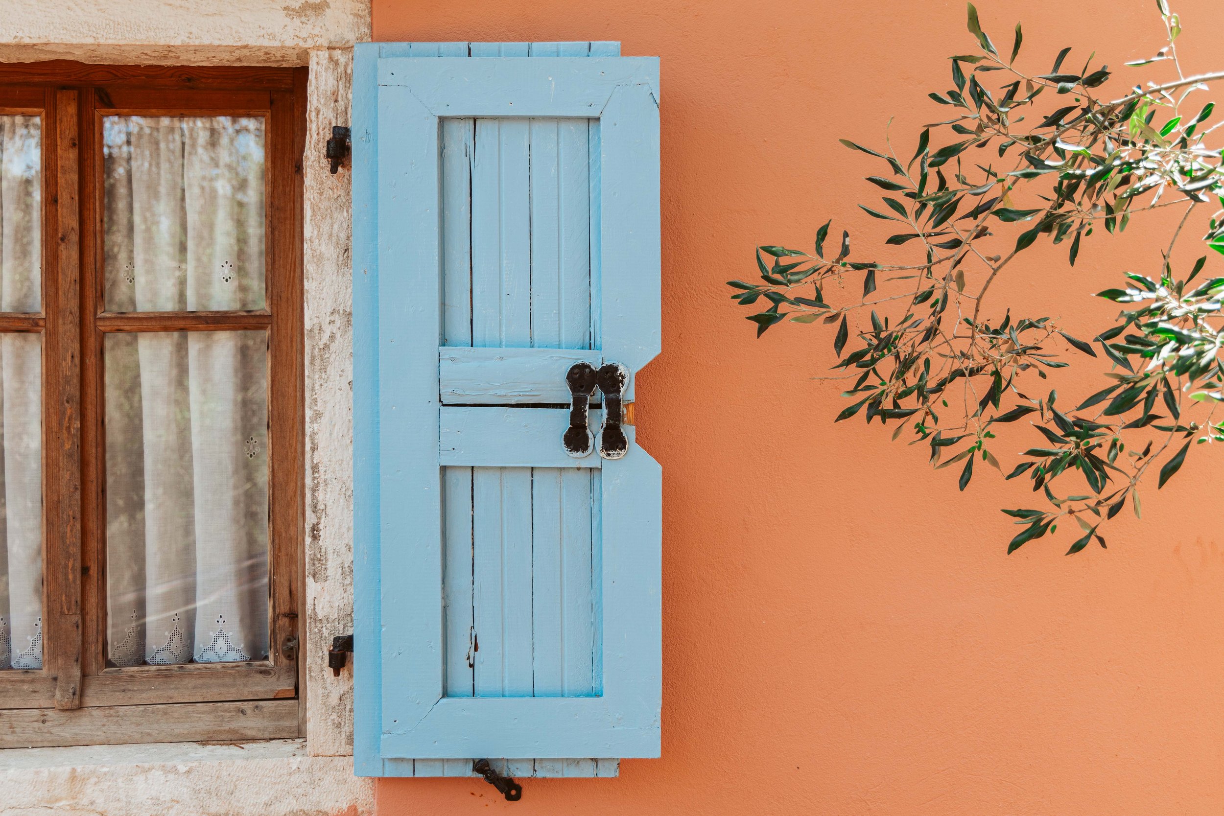 A window with a wooden frame and curtain on a weathered wall, next to a light blue wooden shutter on a peach-colored wall, with a branch of an olive tree on the right.