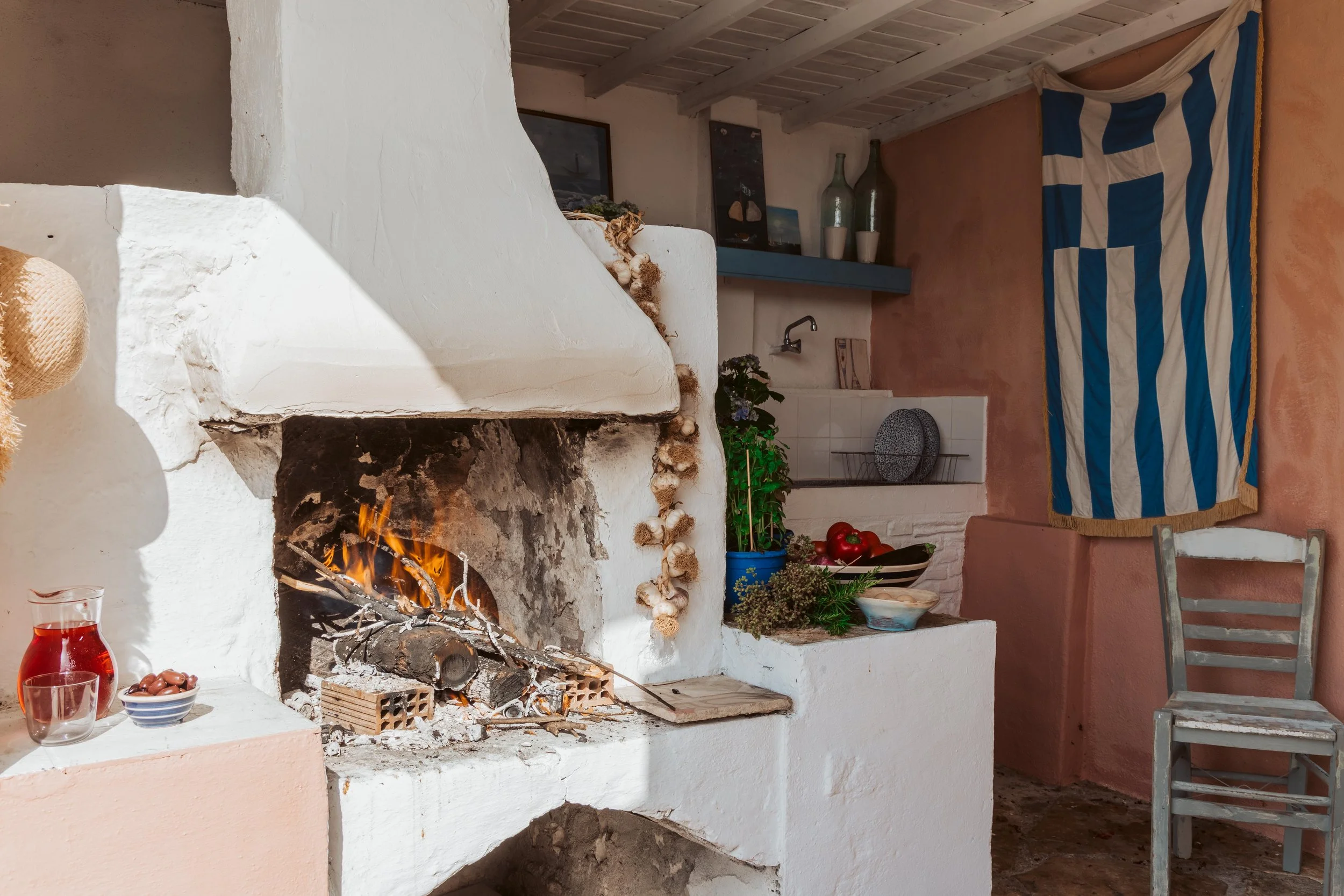 Cozy indoor scene featuring a white stucco fireplace with a wood fire, decorated with garlic braids, adjacent shelves with blue and white plates, green plants, bowls of food, a Greek flag hanging on the wall, and a wooden chair.