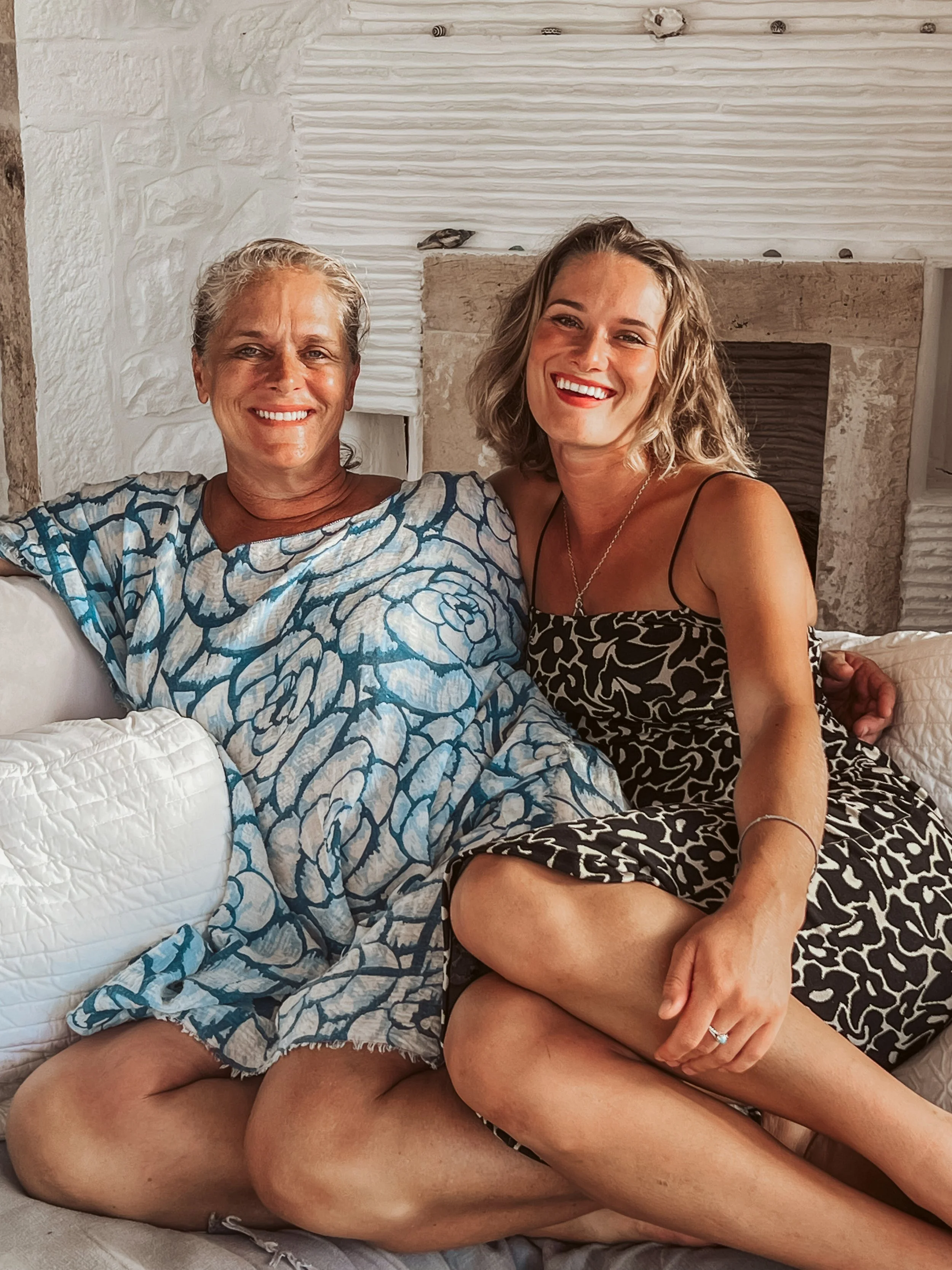 Two women sitting on a white couch, smiling. The woman on the left has short gray hair and is wearing a blue and white patterned dress. The woman on the right has shoulder-length blonde hair and is wearing a black and white patterned sleeveless dress