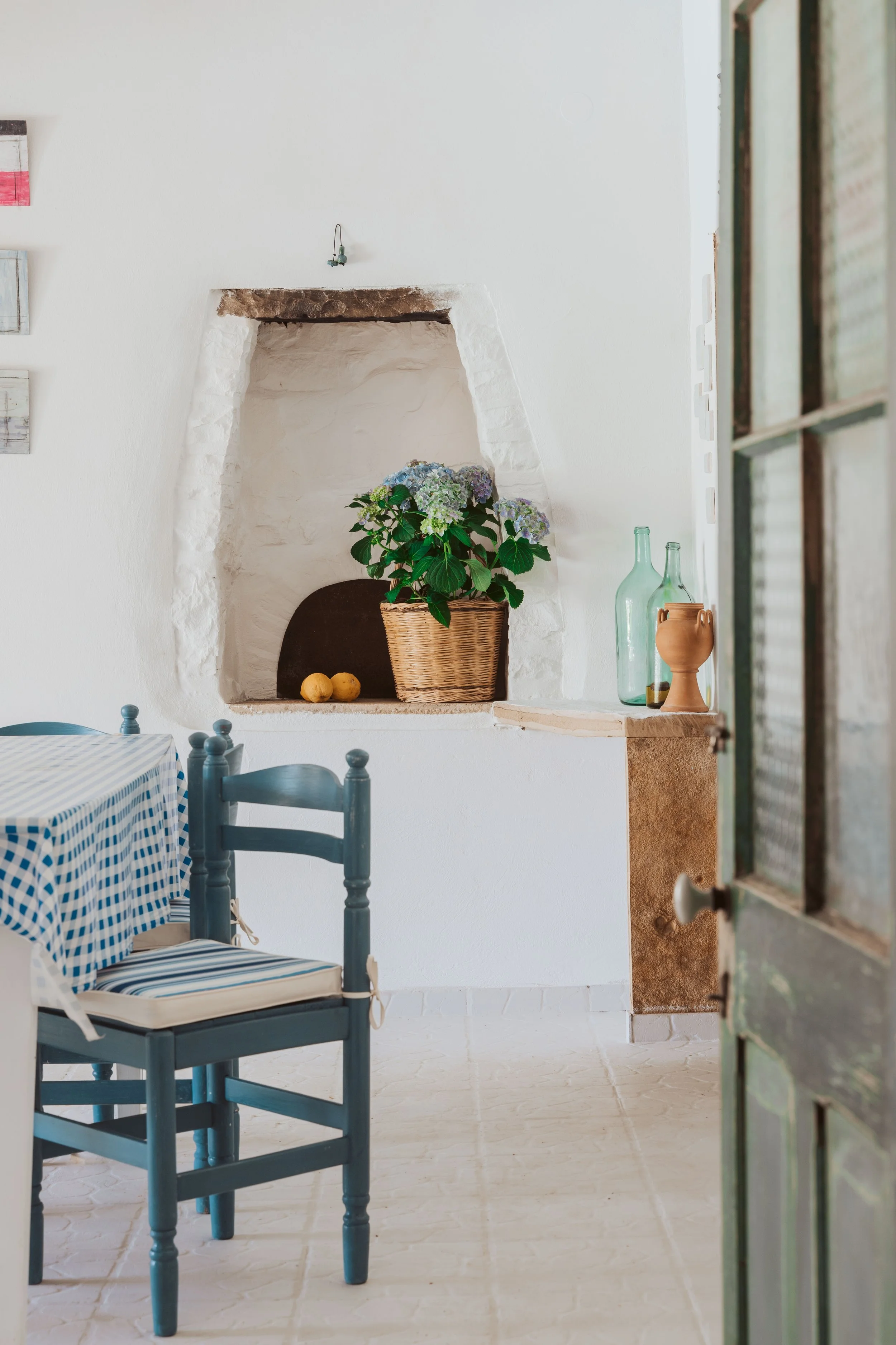 A cozy dining area with a blue checkered tablecloth on the table, blue wooden chairs, a white textured wall with a niche holding a potted hydrangea plant, glass bottles, and pottery, a tiled floor, and a partially open rustic green door.
