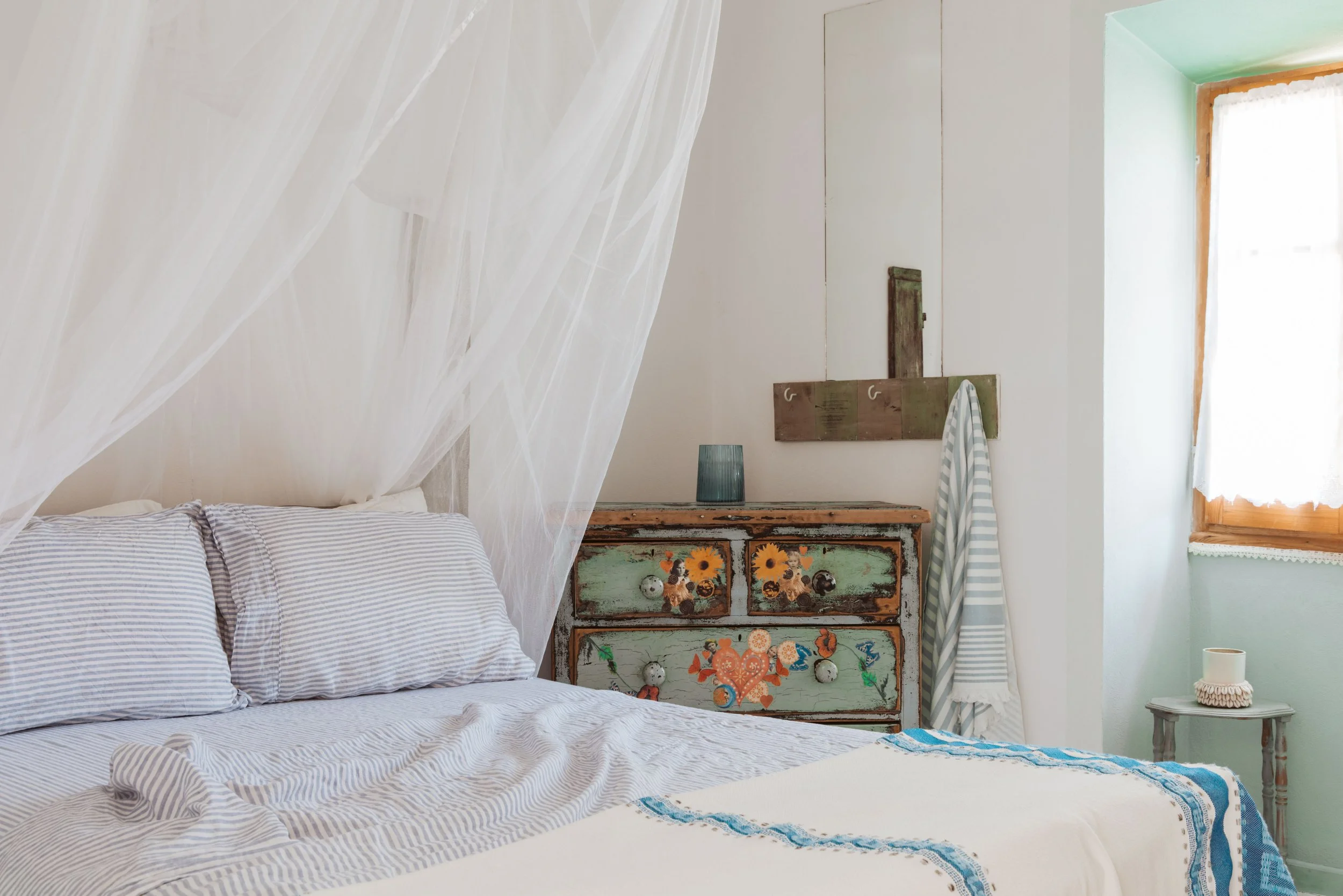 Cozy bedroom with striped bedding, an antique painted dresser with floral designs, a striped towel hanging on a hook, a small white side table with a candle, and a window with light curtains.