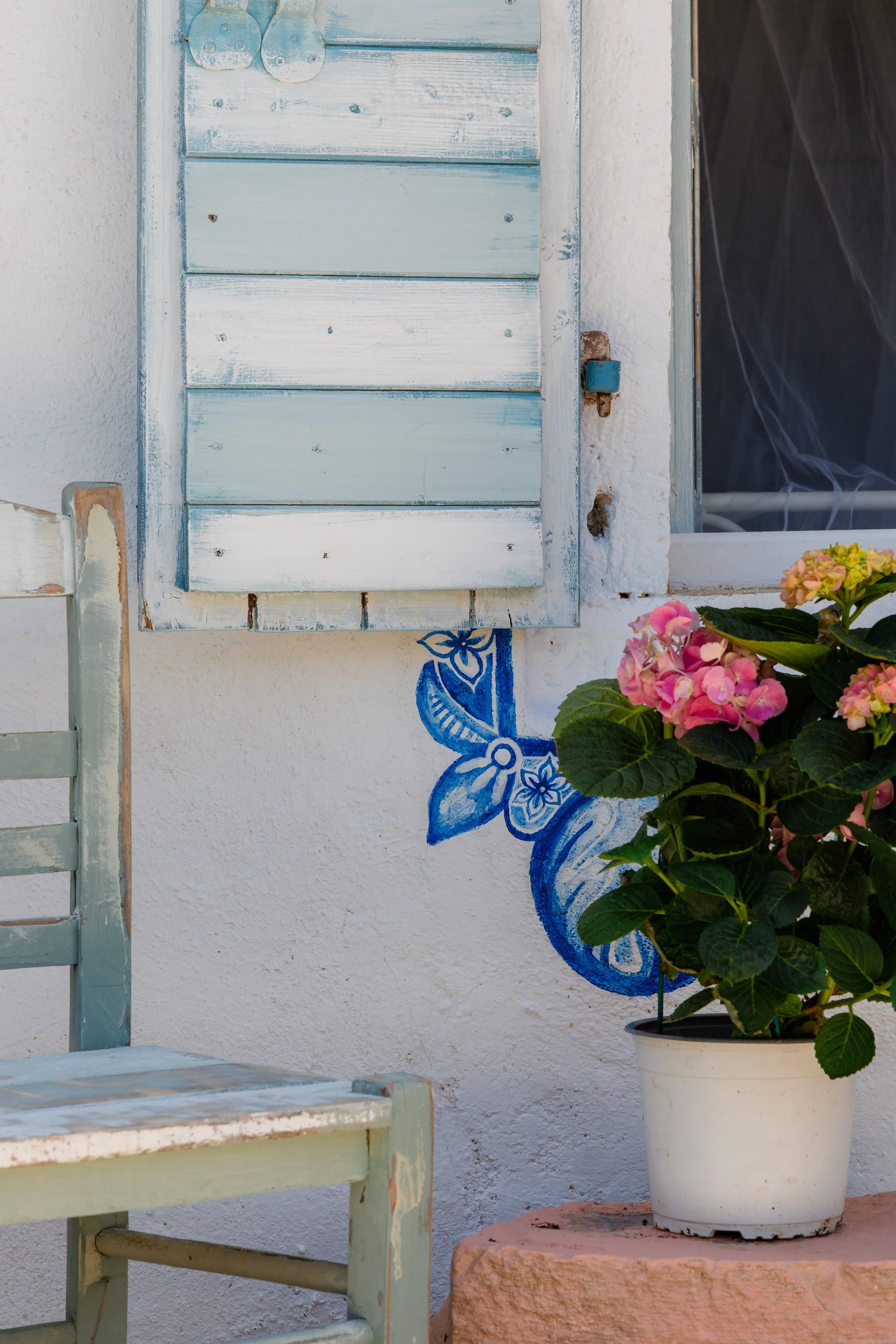 A white wall with a painted blue flower decorative design, green wooden chairs, a potted plant with pink hydrangea blossoms, and a light blue window shutter.