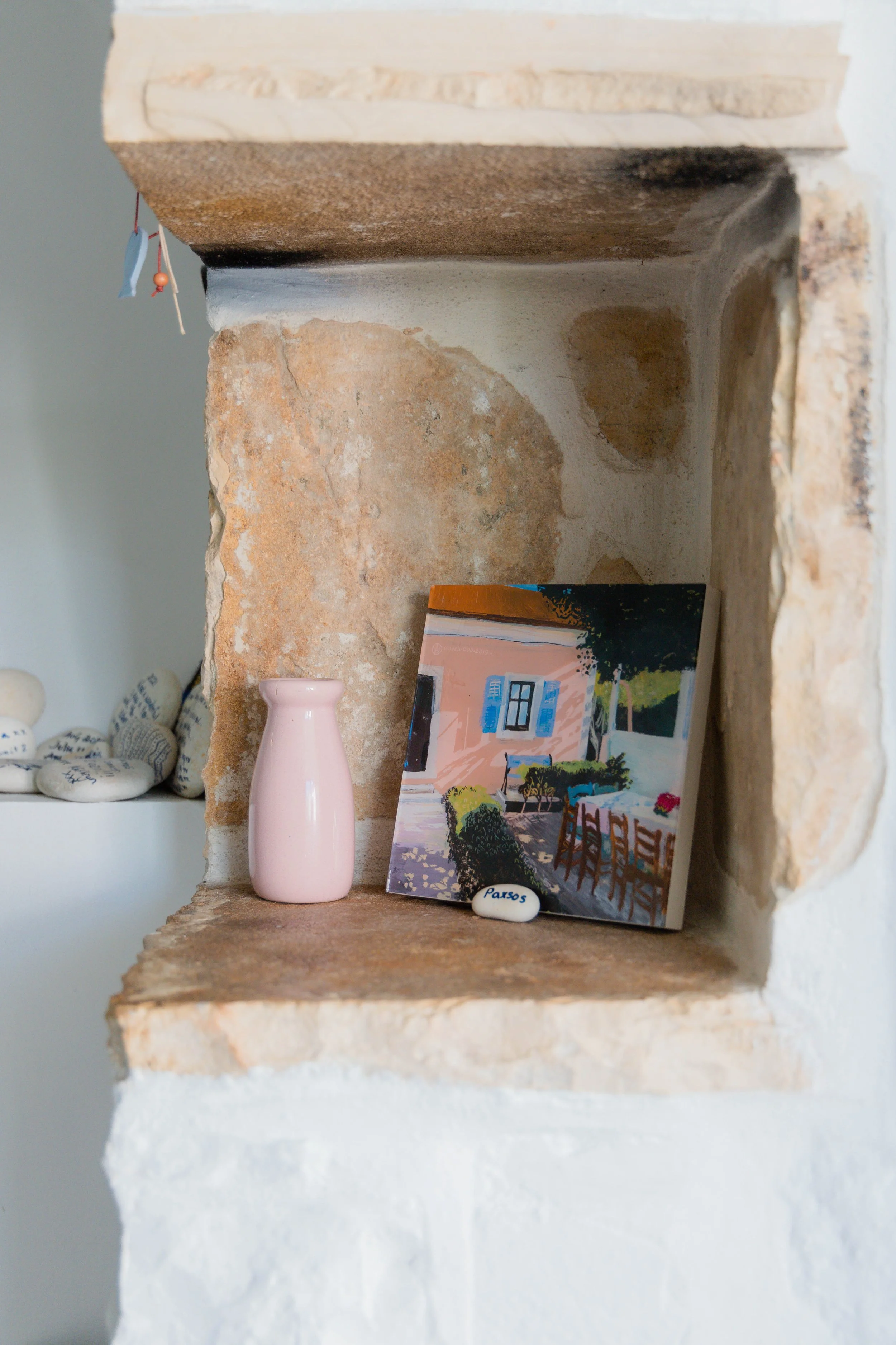 Decorative shelf with a pink vase, a landscape painting, and a small stone with the word "Paros" written on it, against a rustic stone wall.