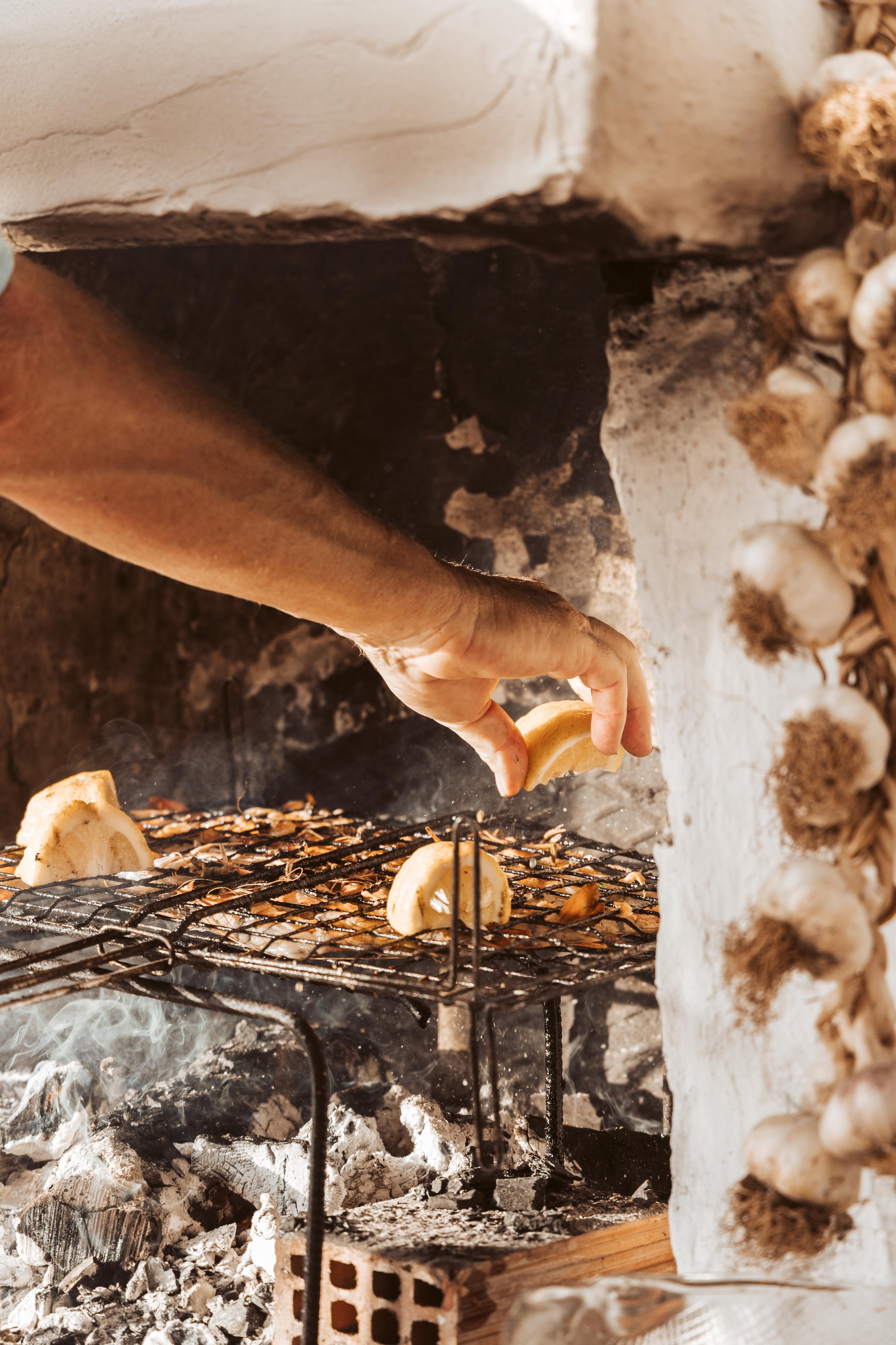 Person placing banana slices on a grill inside a wood-fired oven, with garlic bulbs roasting on the side.