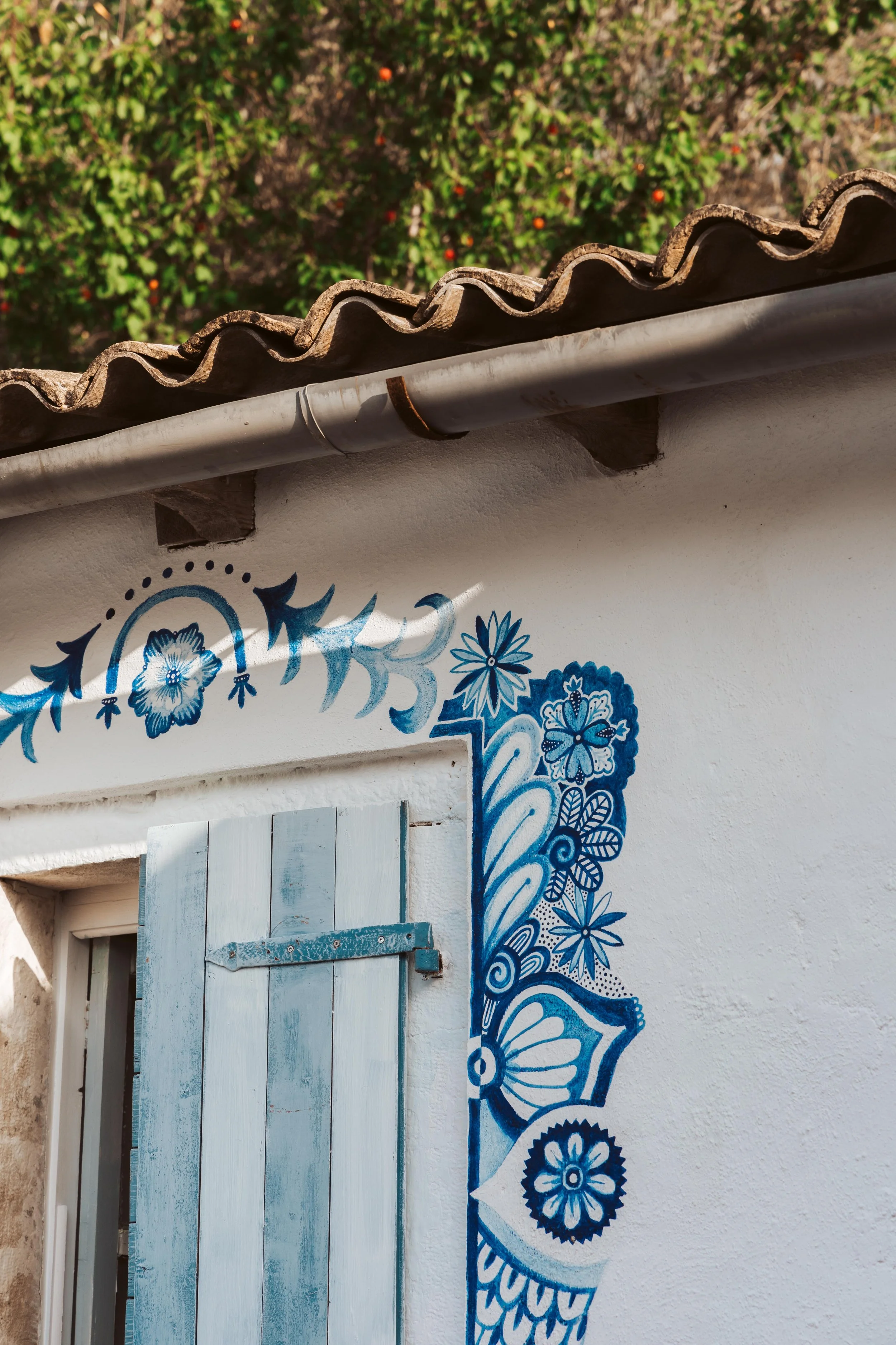 A white wall with blue floral and butterfly artwork framing a small window with blue wooden shutters, a tiled roof, and a bushy tree in the background.