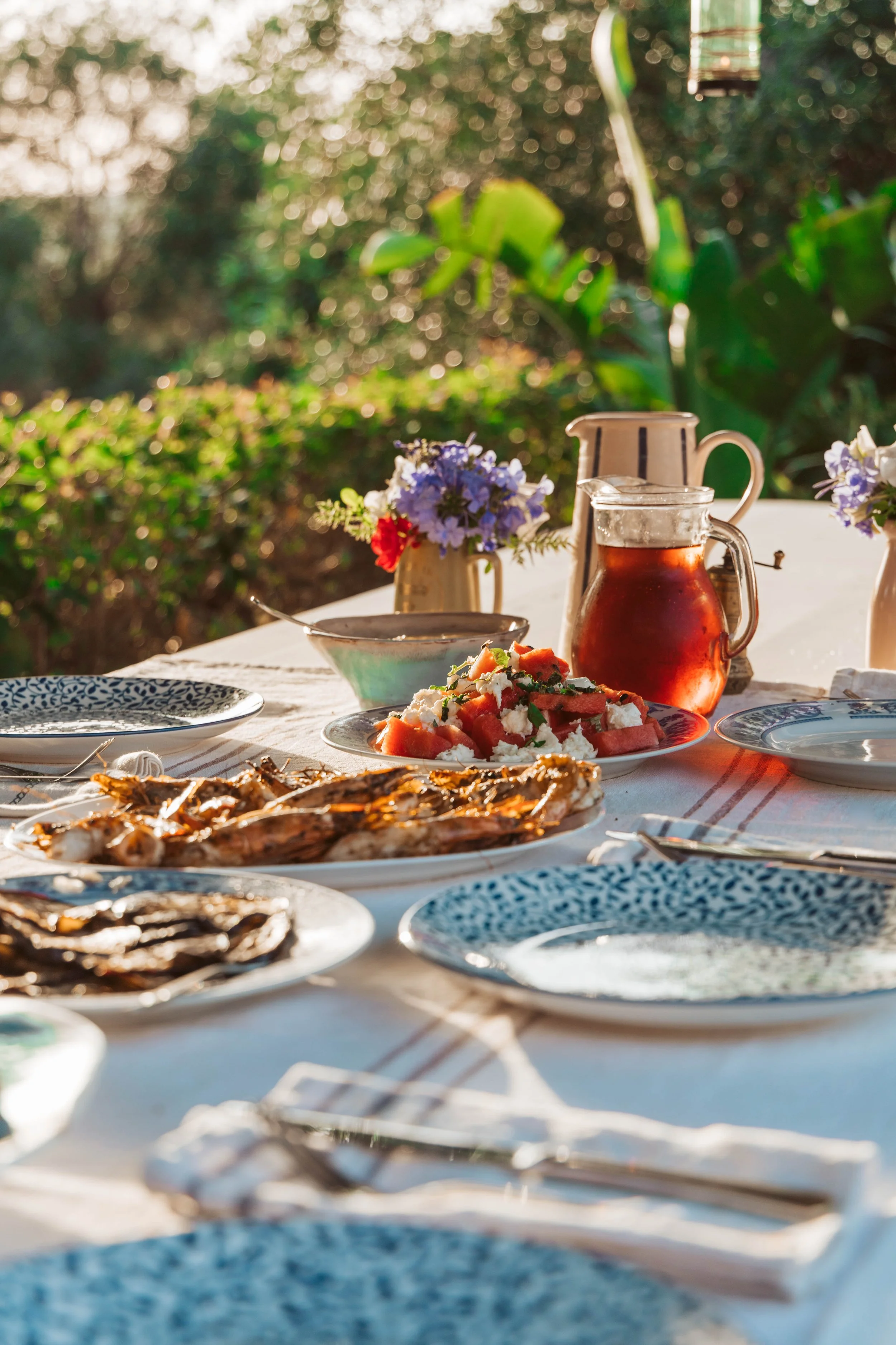 Outdoor dining table set with plates of food including grilled meats, watermelon salad, and a pitcher of iced tea, with small vases of flowers, on a sunny day with greenery in the background.