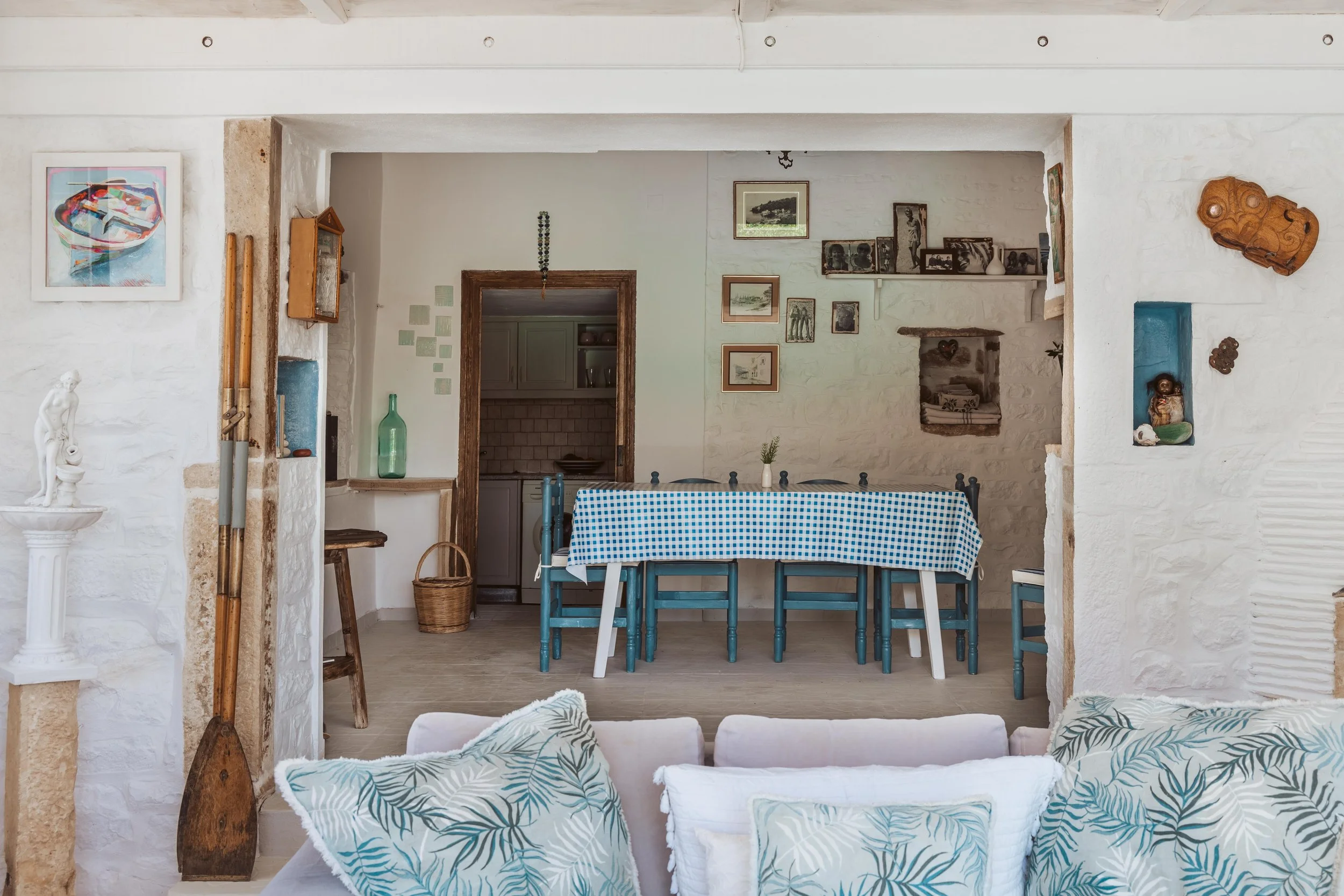 A cozy indoor dining area with a long table covered with a blue checkered tablecloth and surrounded by blue and white chairs. The room features white painted brick walls, recessed shelves displaying decorative items, and a collection of framed pictur