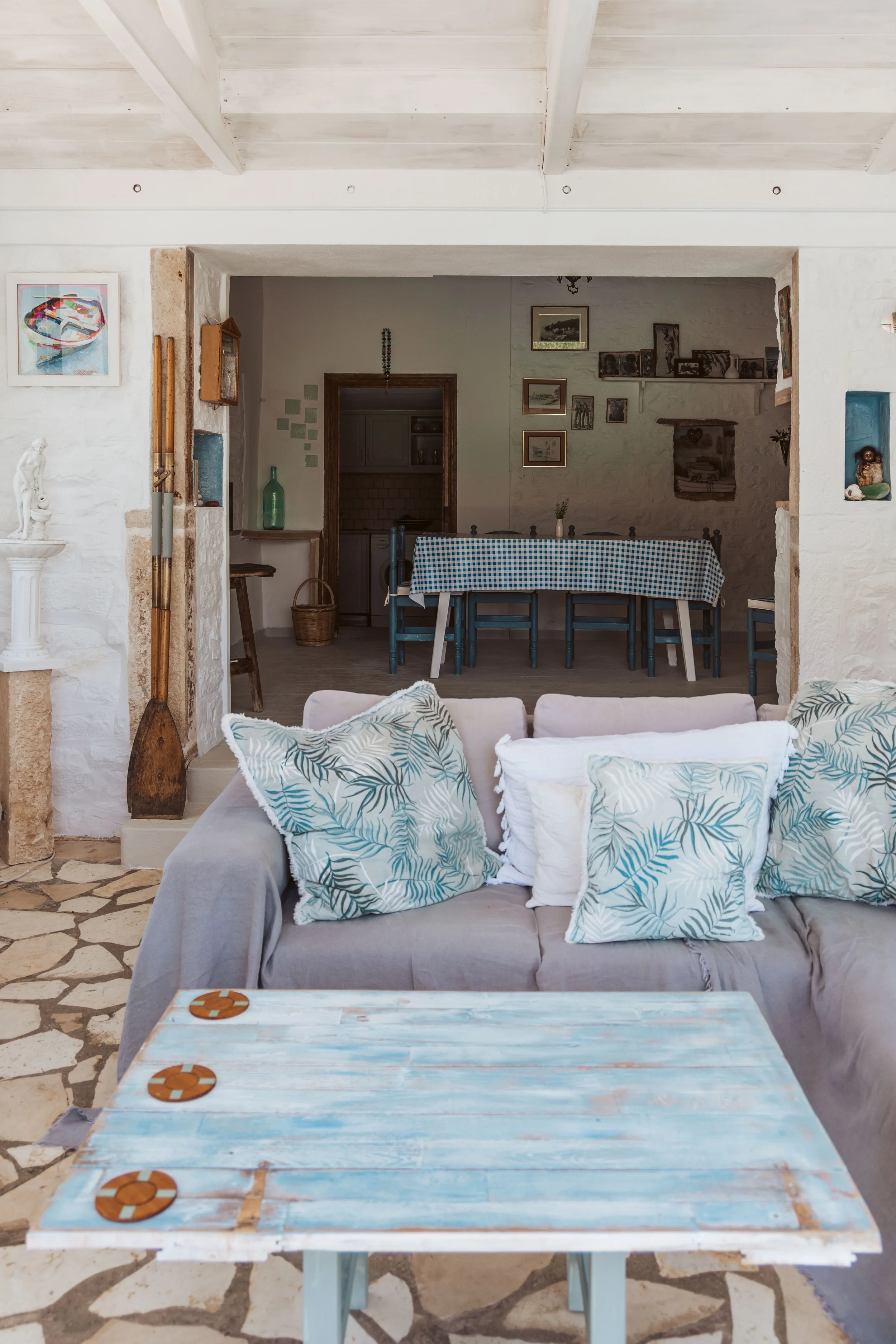 Living room with a beige sofa and blue patterned cushions, a distressed wooden coffee table with orange sauce containers, stone flooring, and an open dining area with a checkered tablecloth.