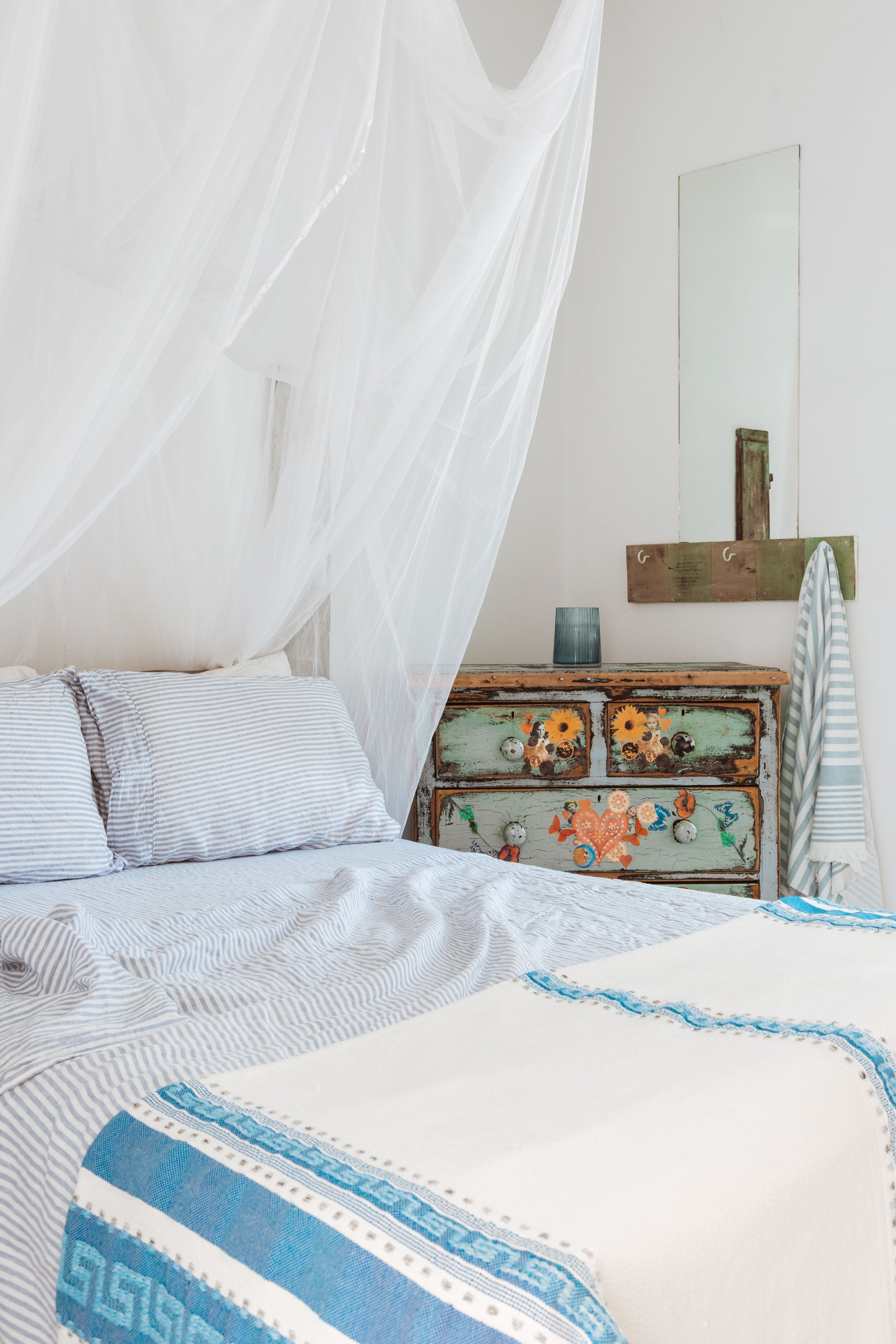 A cozy bedroom with a white bed, blue and white striped pillows, and a sheer white canopy. A rustic wooden dresser with floral and heart motifs is beside the bed, with a small blue vase and a mirror on the wall behind.
