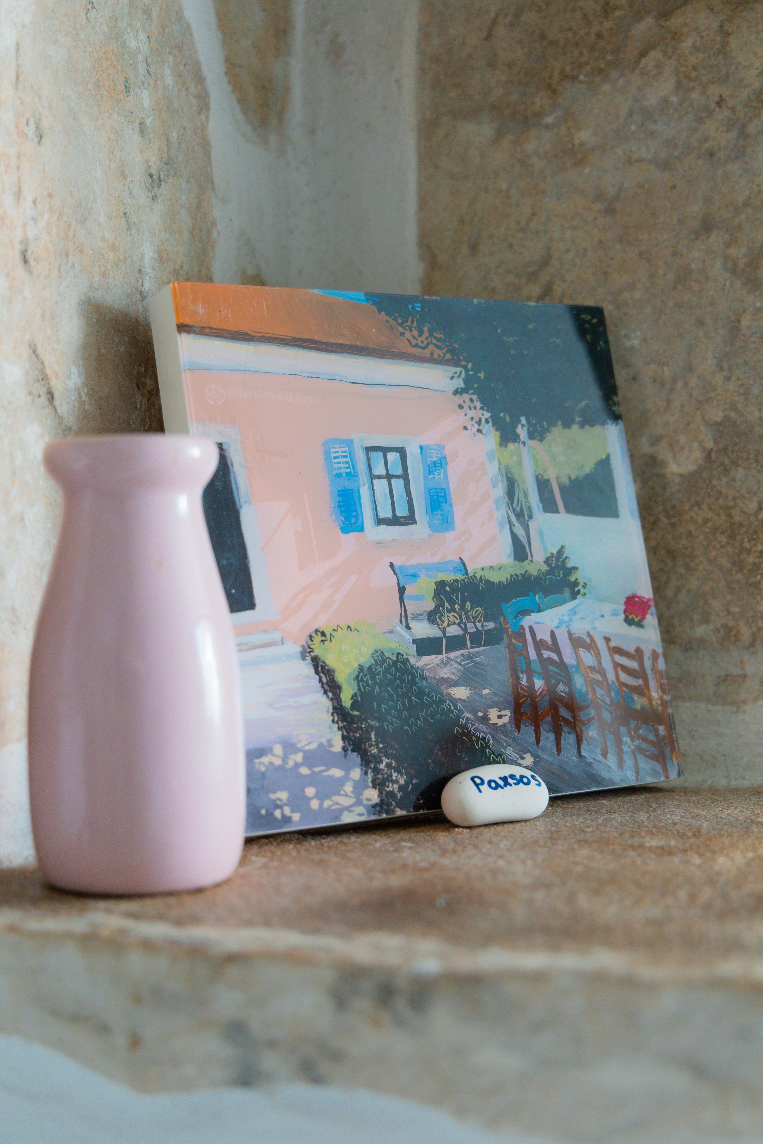 A painted canvas of a house with a porch, chairs, and bushes, a small pink vase, and a white stone with the word 'Paxos' written on it, placed on a brick ledge against a brick wall.
