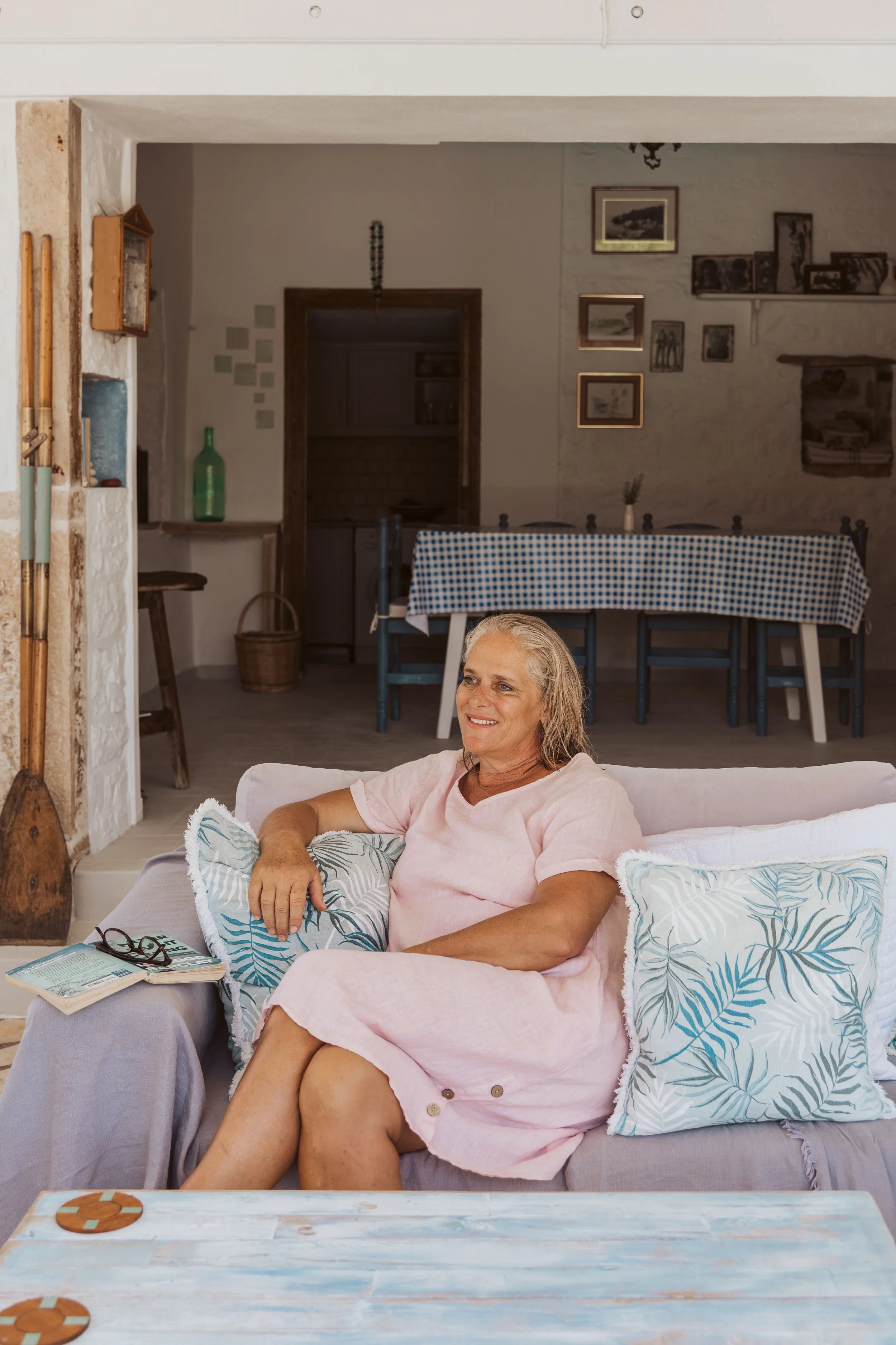 A woman with gray hair sitting on a sofa, smiling, wearing a light pink dress, holding a pillow, with glasses, a book, and a coaster on the side table.