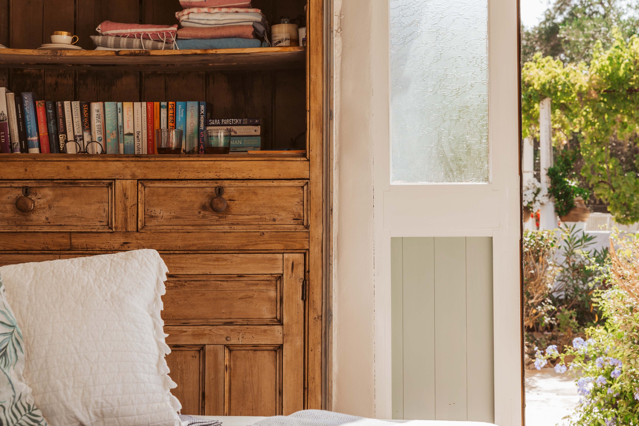 A cozy bedroom with a wooden bookshelf filled with books and towels, next to a glass door leading to a garden with greenery and blooming flowers.