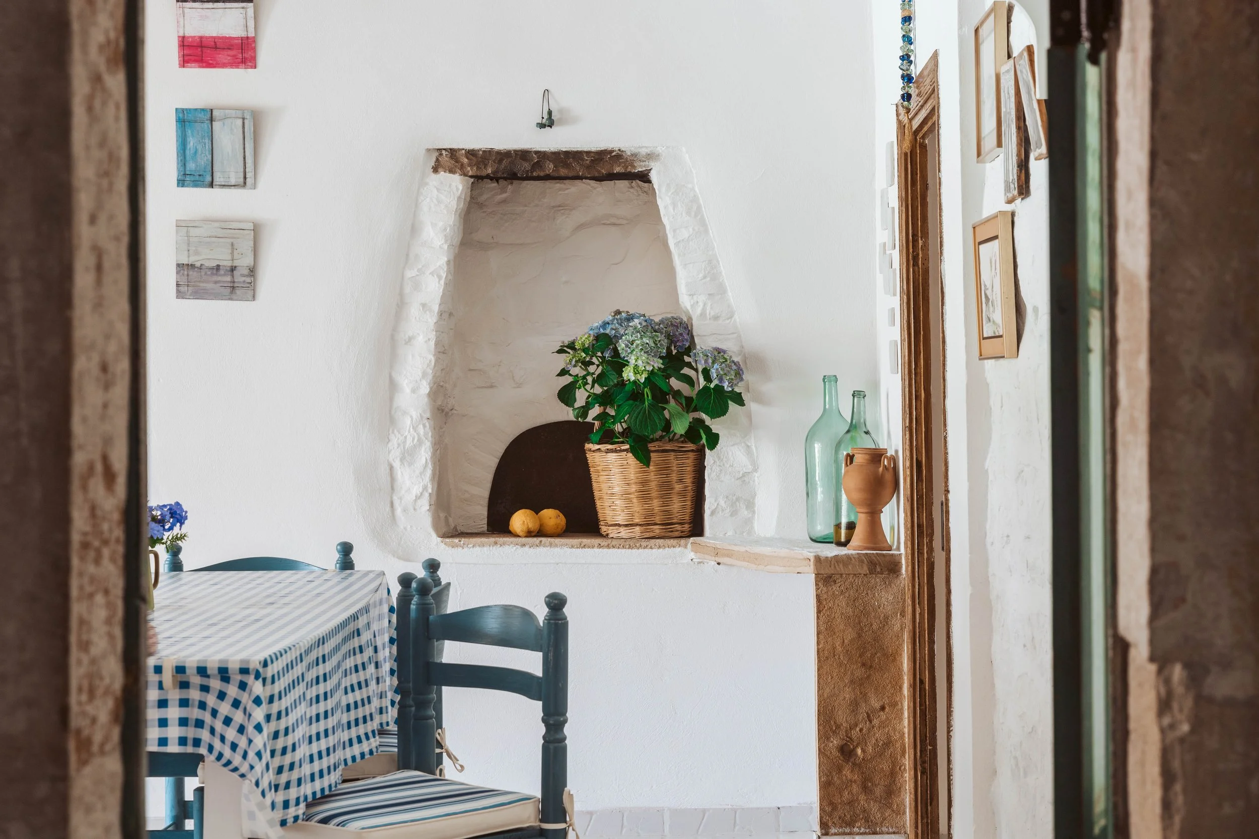 Interior of a cozy kitchen or dining area with a white wall, a built-in niche with a flower pot and lemons, a table with a blue and white checkered tablecloth, framed artwork on the wall, and decorative bottles and pottery on a shelf, seen from a doo