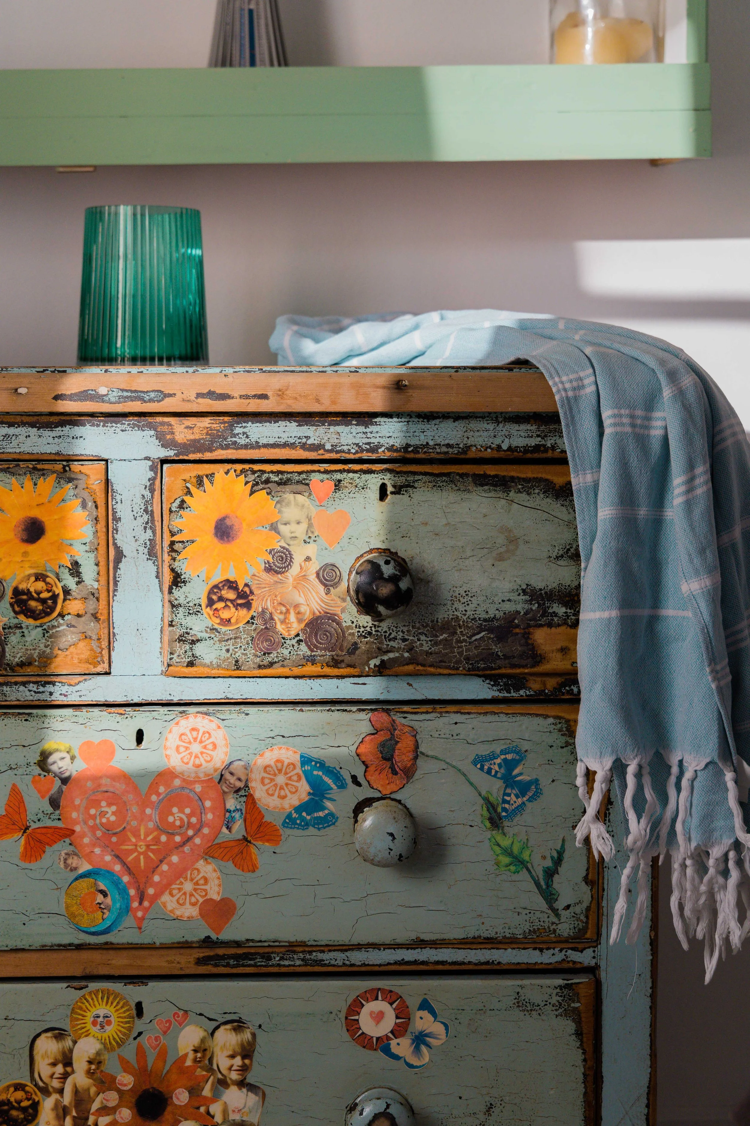 A vintage distressed dresser decorated with colorful paper cutouts of flowers, faces, and butterflies. A light blue and white striped cloth hangs over the right side of the dresser. On top of the dresser, there is a green glass lamp and a curtain wit