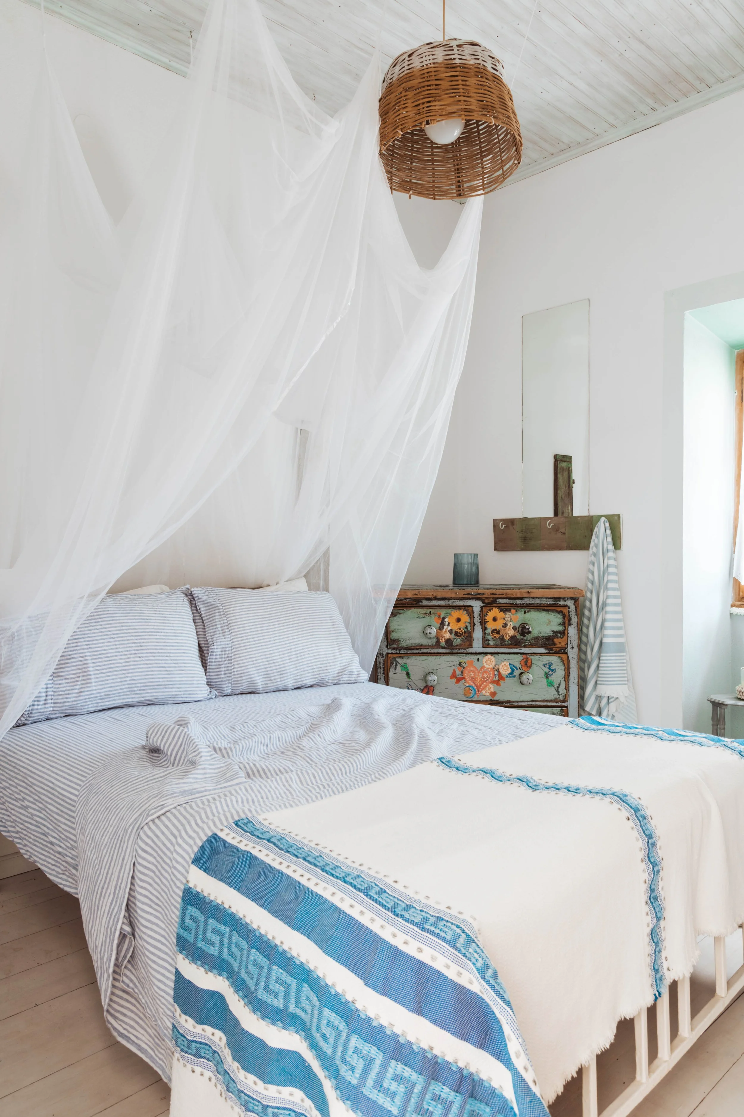 A cozy bedroom with white walls, a bed with gray and white striped bedding, and a blue and white patterned quilt. There is a sheer, white canopy hanging above the bed. To the right is a distressed painted wooden dresser with sunflowers and floral des