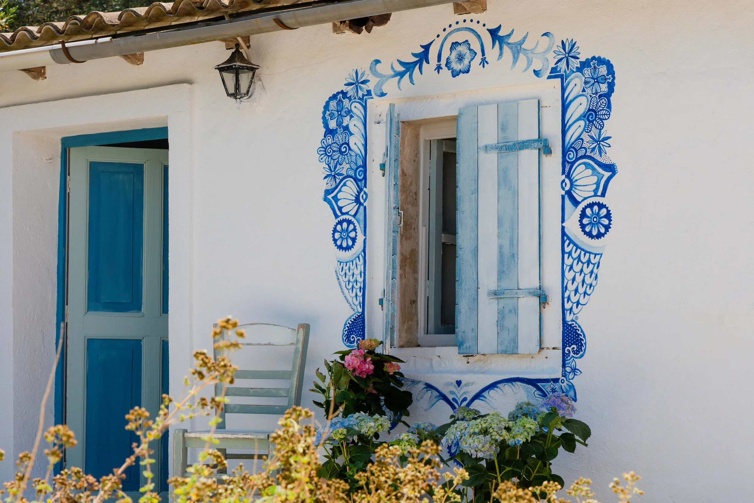 White house with blue window shutters and decorative blue floral and butterfly patterns around the window. There are flowers and plants in front, and an outdoor chair beside the window.