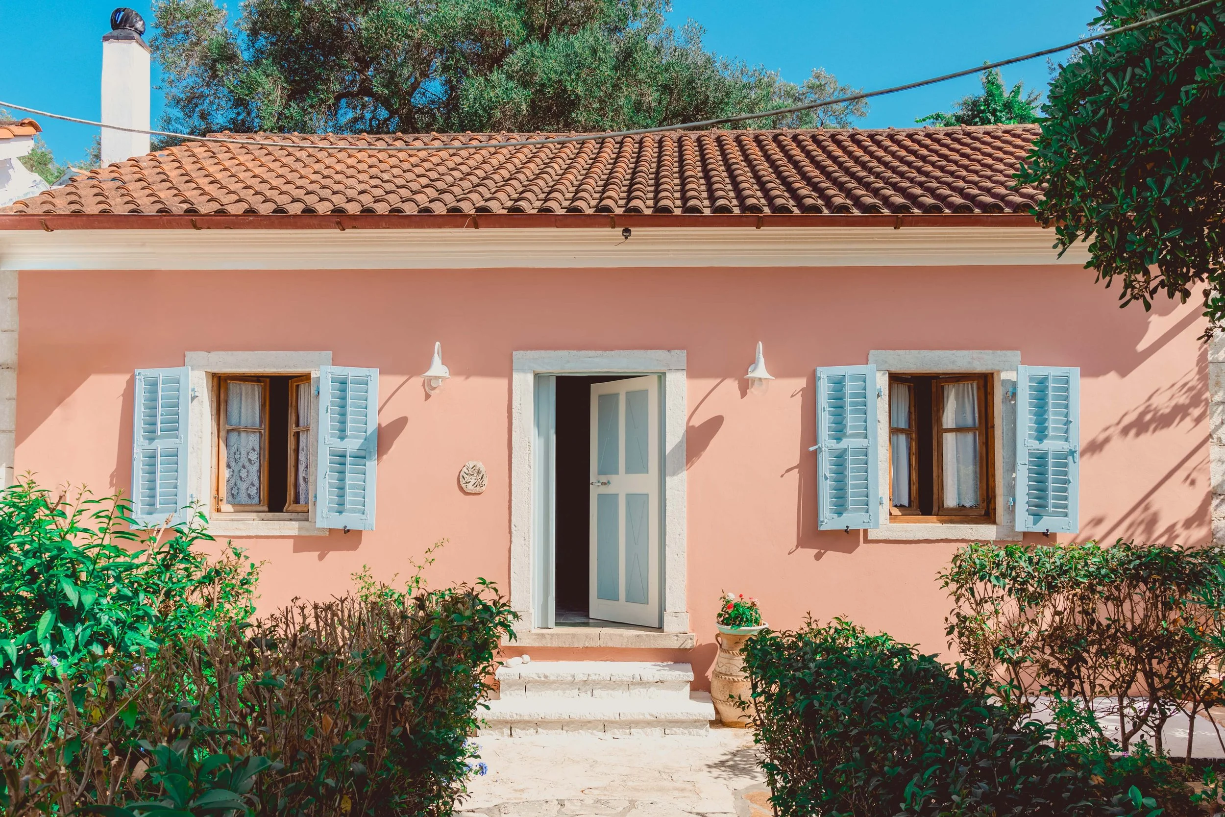 A pink house with a terracotta tile roof, two open windows with light blue shutters, a white door slightly ajar, stone steps leading to the entrance, surrounded by bushes, with a large tree in the background and a clear blue sky.