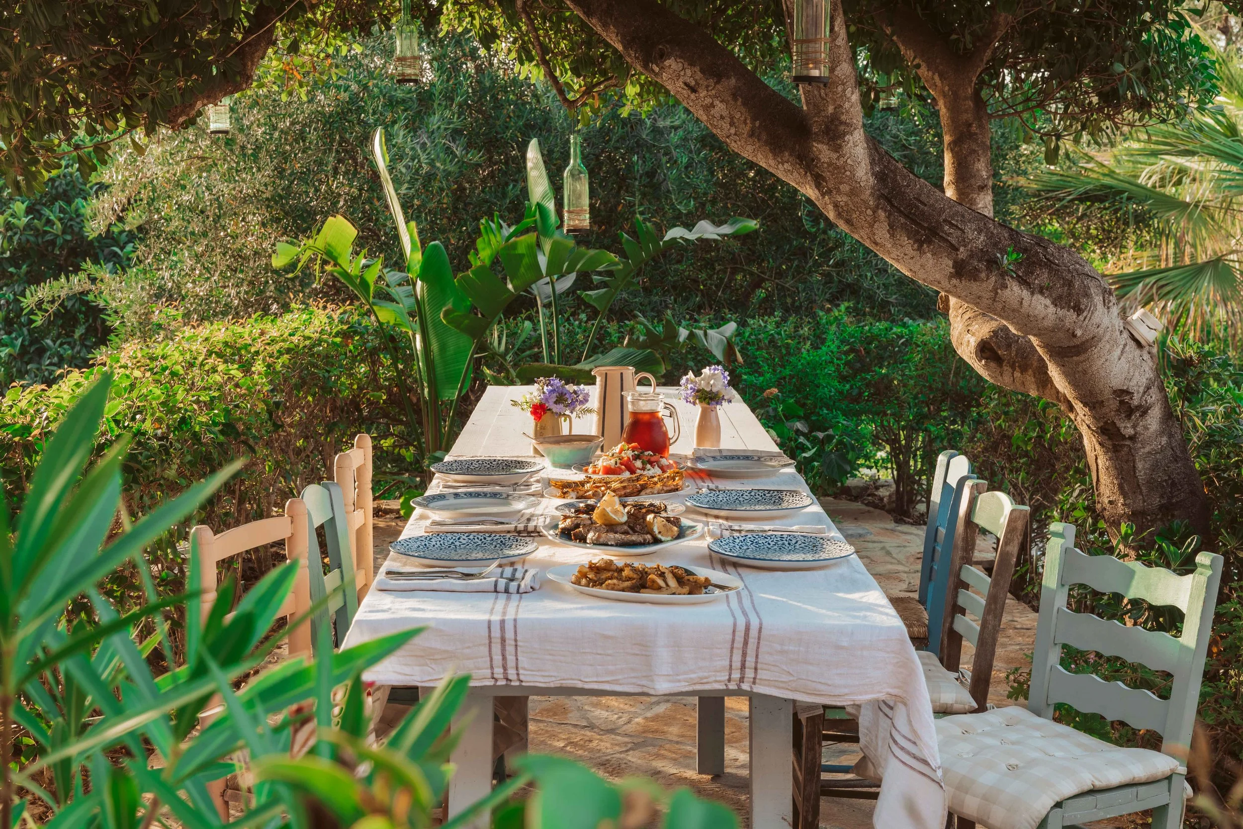 Outdoor dining table set for a meal, with plates, bowls, and pitchers, surrounded by lush green plants and trees.