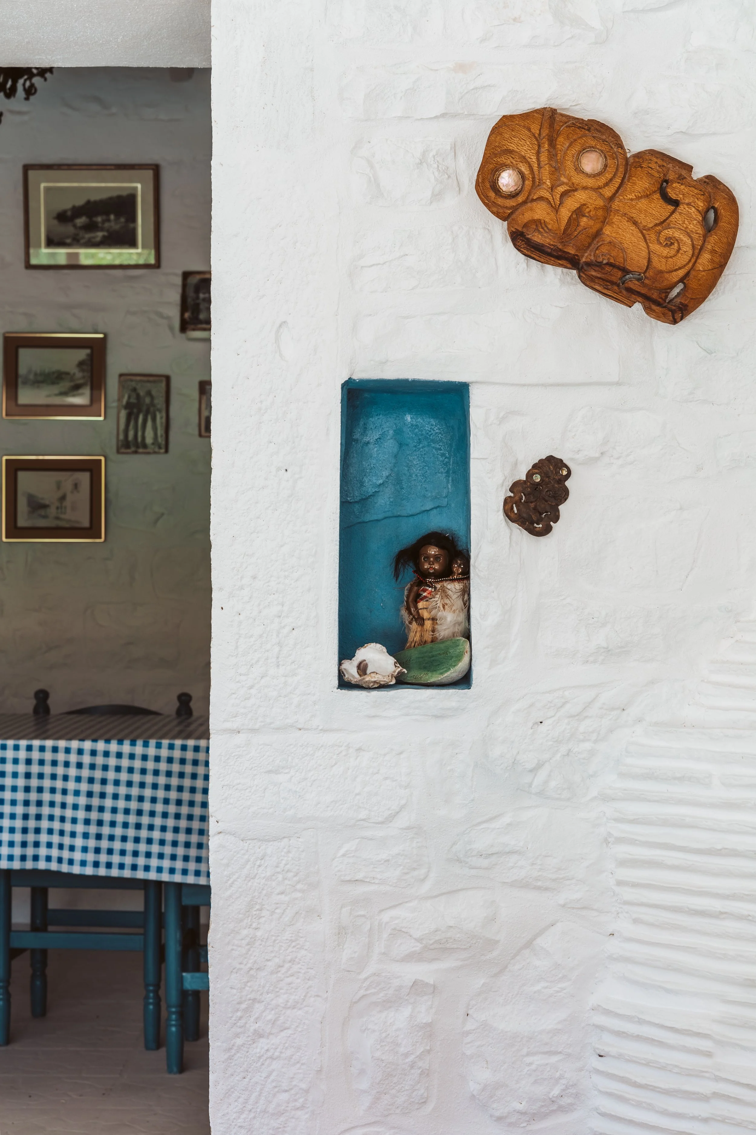 Decorative wooden masks and a doll in a small blue alcove on a white textured wall in a room with photos on the opposite wall and a blue checkered tablecloth.