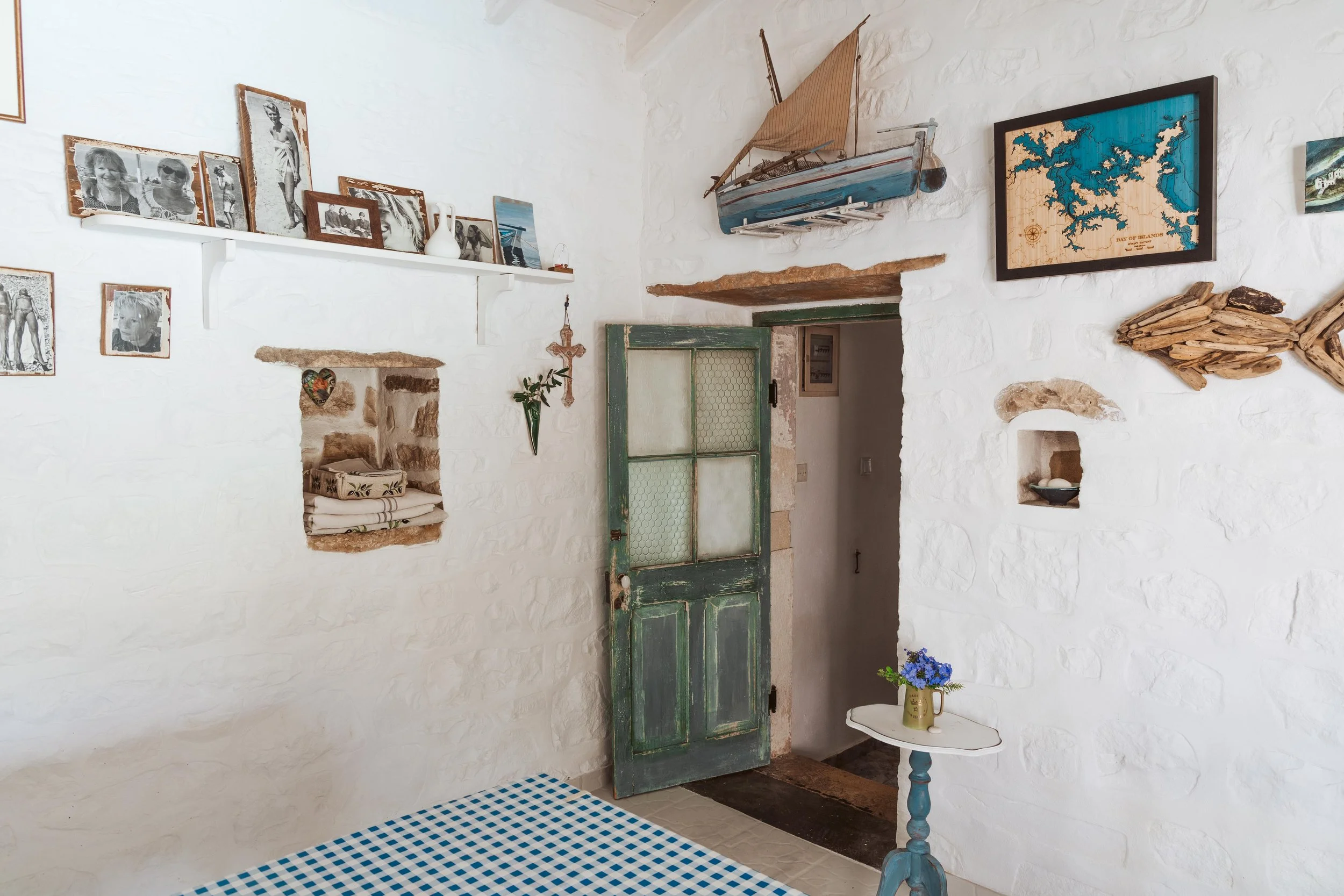 Interior of a rustic room with white textured walls, a green wooden door, and various nautical and vintage decor. There is a model sailboat, framed maps, black and white photos, and a small table with a flower vase.