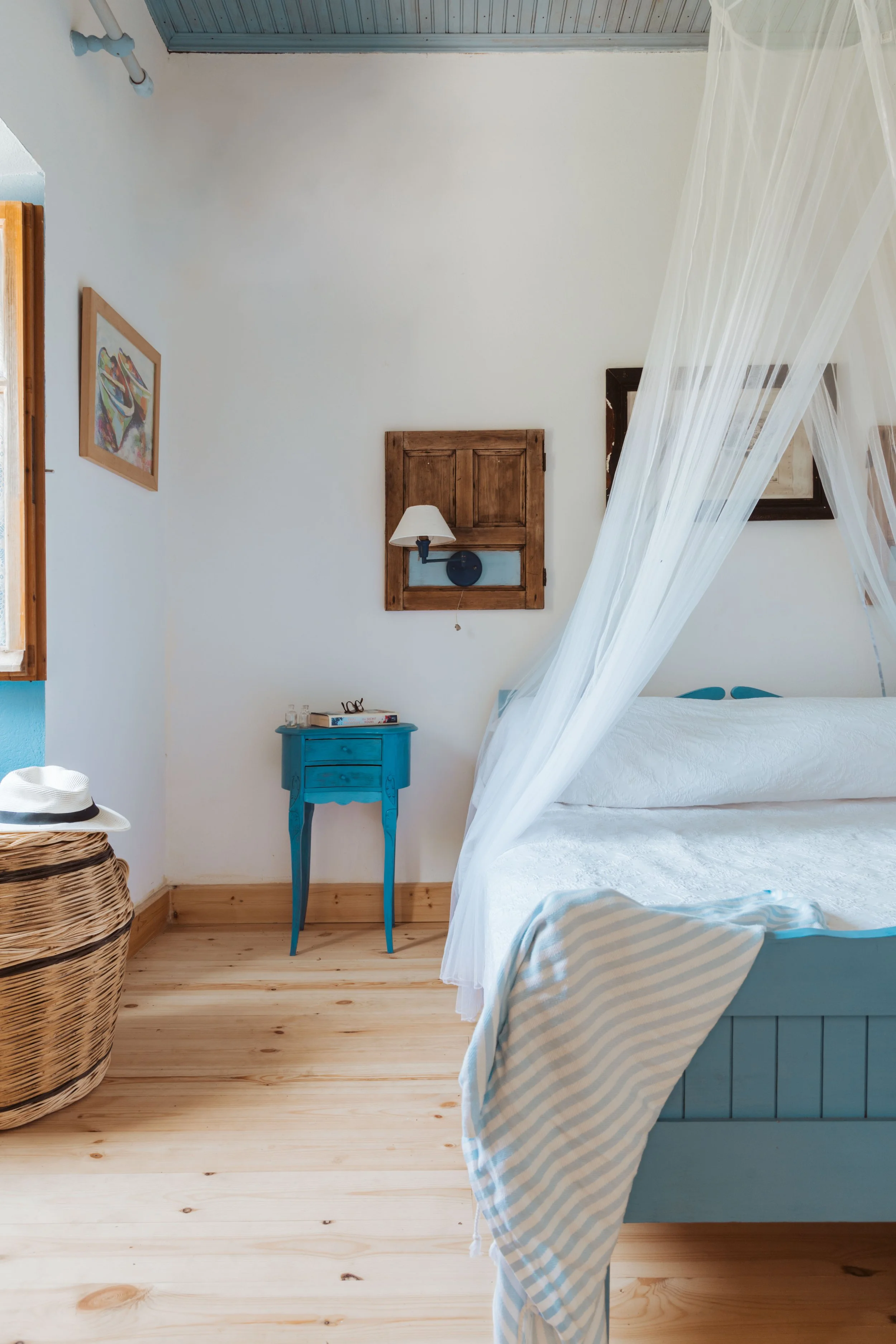 A bedroom with a white bed, sheer canopy, blue bedside table, wooden floors, and wall decor, including a picture and a wall-mounted lamp, window with wooden trim, and a wicker basket with a white hat.