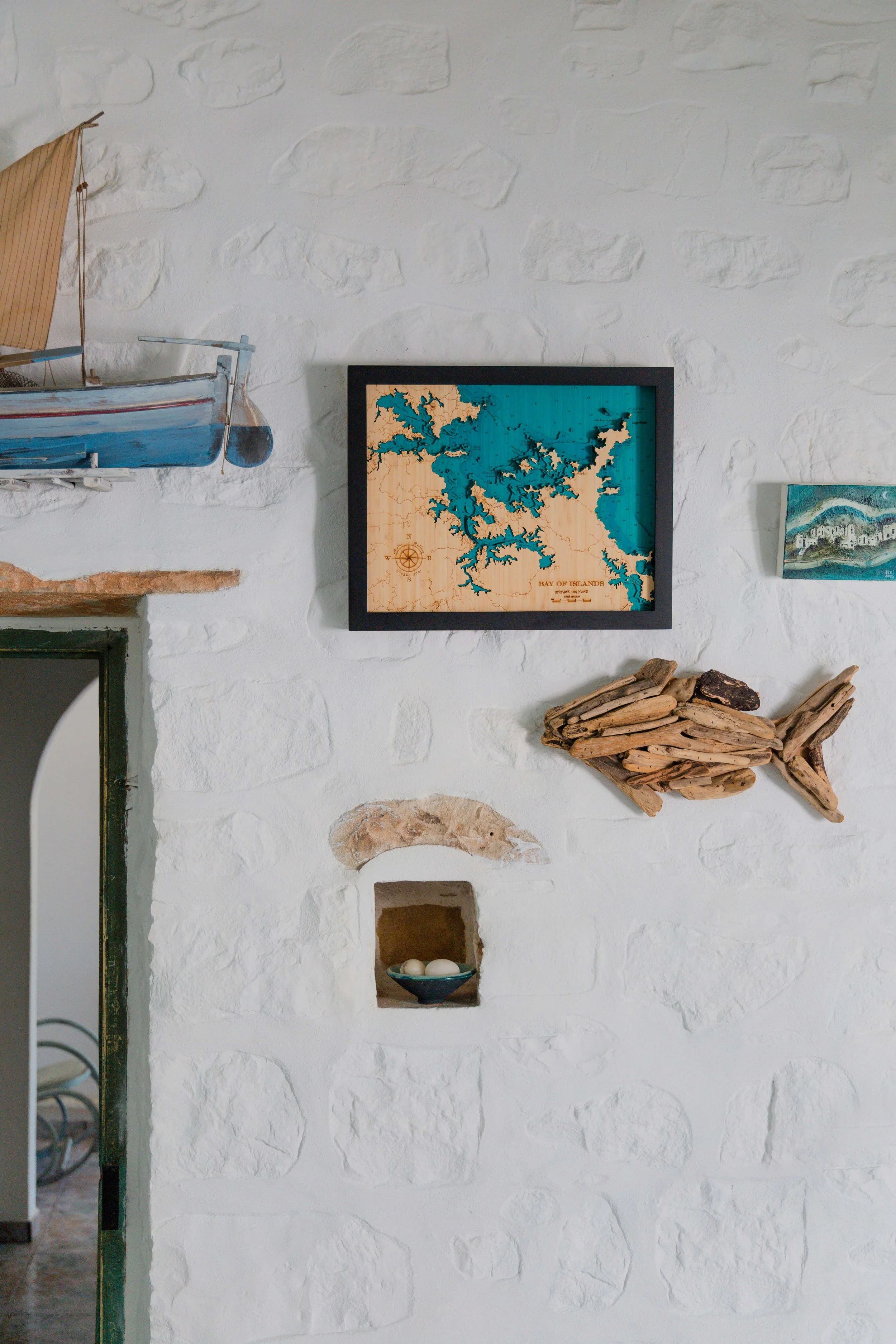 Decorative wall with model sailboat, framed map of Bay of Islands, small landscape painting, driftwood fish sculpture, and a small niche with eggs in a bowl.