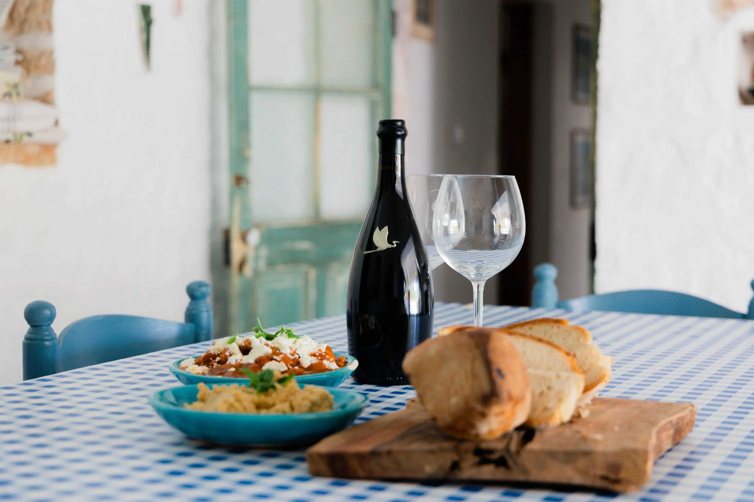 Table set with bread, two blue bowls of food, a bottle of red wine, and empty wine glasses on a checkered tablecloth in a rustic room.