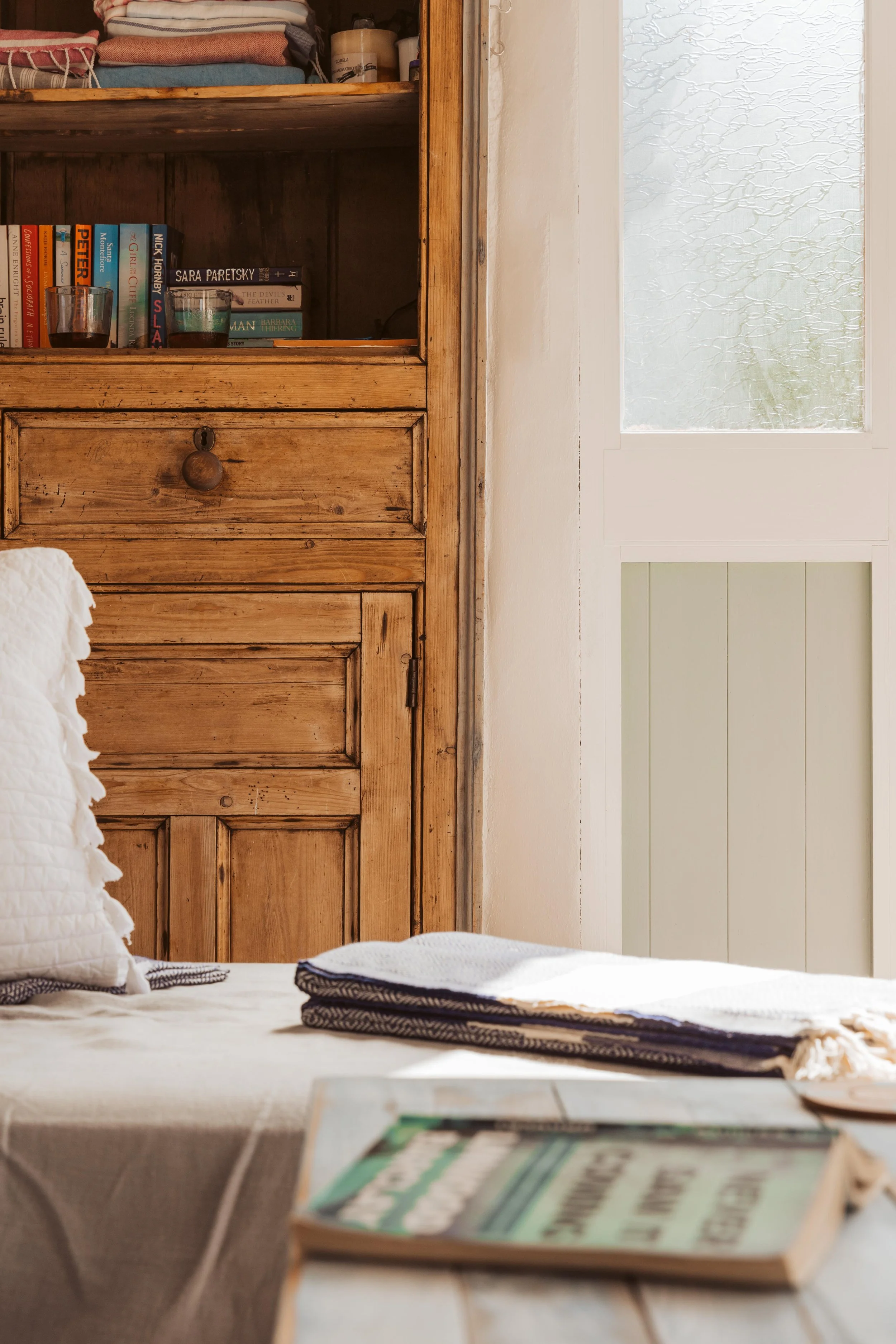 A cozy room with a wooden cabinet, a white door with glass, a bed with white pillow and blanket, and a book on the table near the bed.