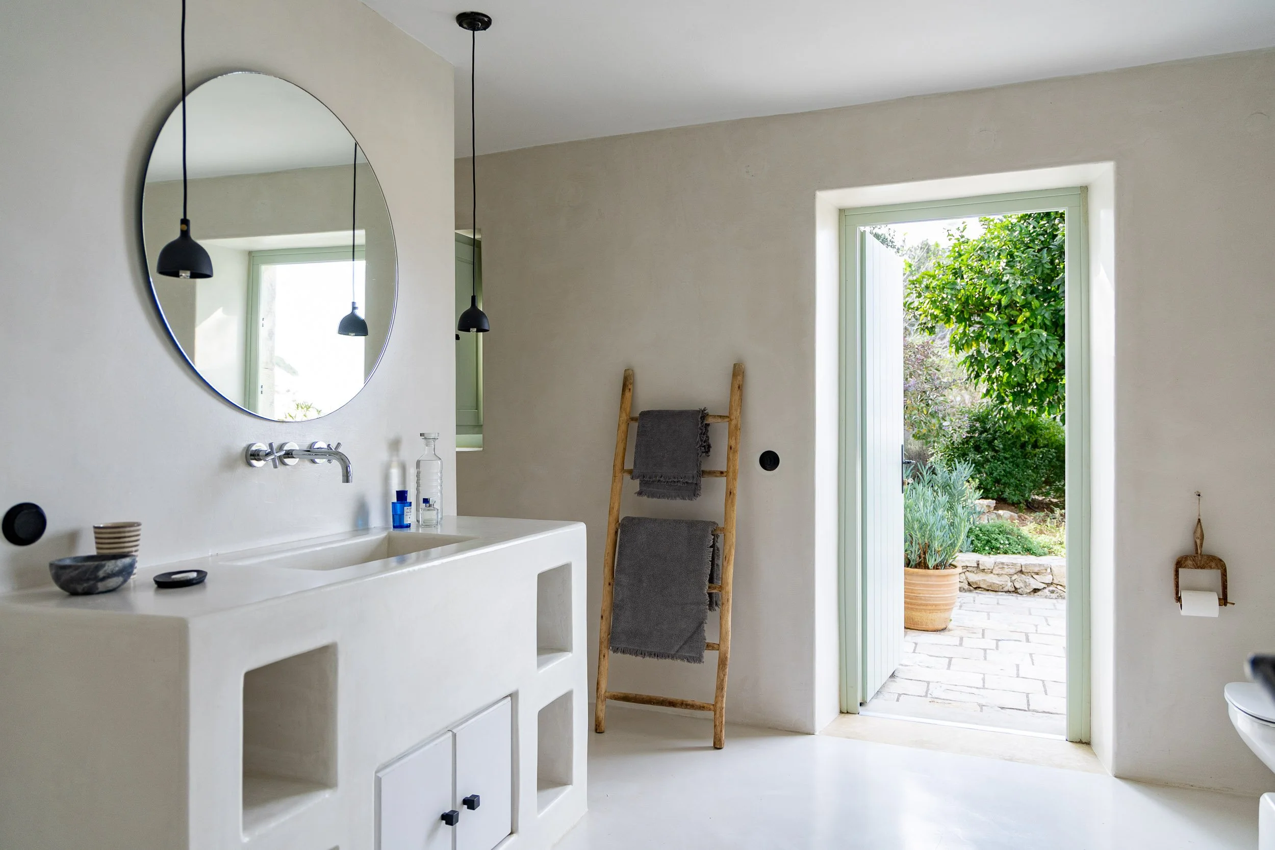 Modern bathroom with white sink, round mirror, black pendant lights, wooden towel ladder with gray towels, open door leading to garden, and minimal decor.