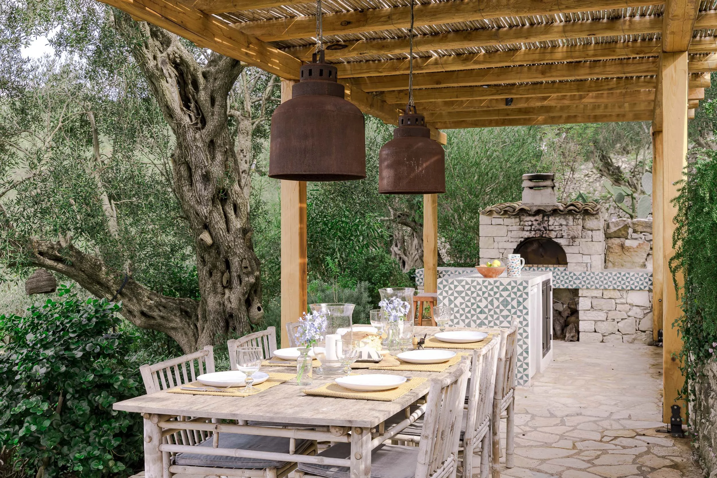 Outdoor patio dining area with a rustic wooden table set for a meal, surrounded by wooden chairs, under a patio cover with two large hanging lamps. In the background, there is a stone oven, a counter with decorated tiles, and lush greenery with trees