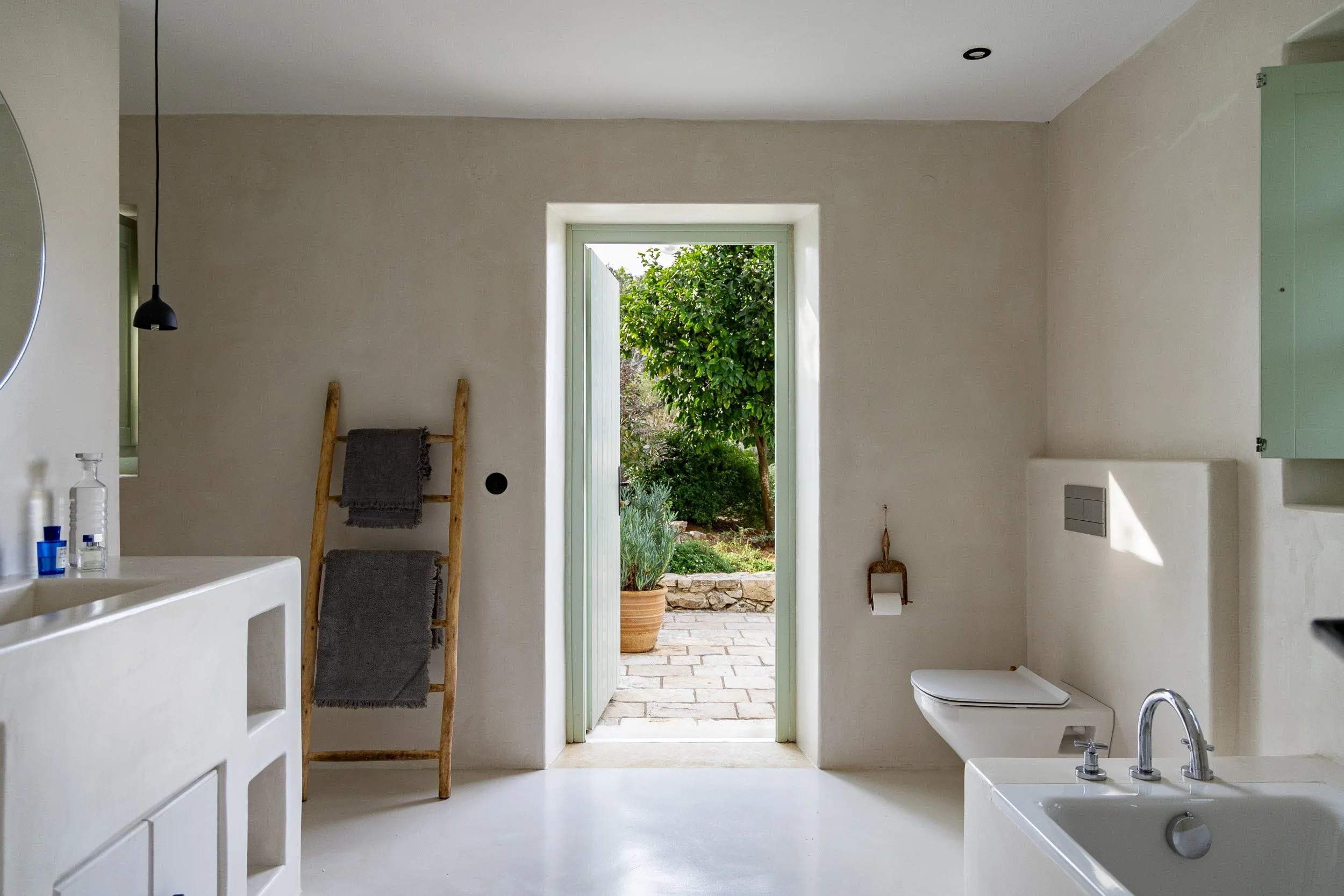 Modern minimalist bathroom with white walls and fixtures, a small window, and an open door leading to a garden with greenery and potted plants.
