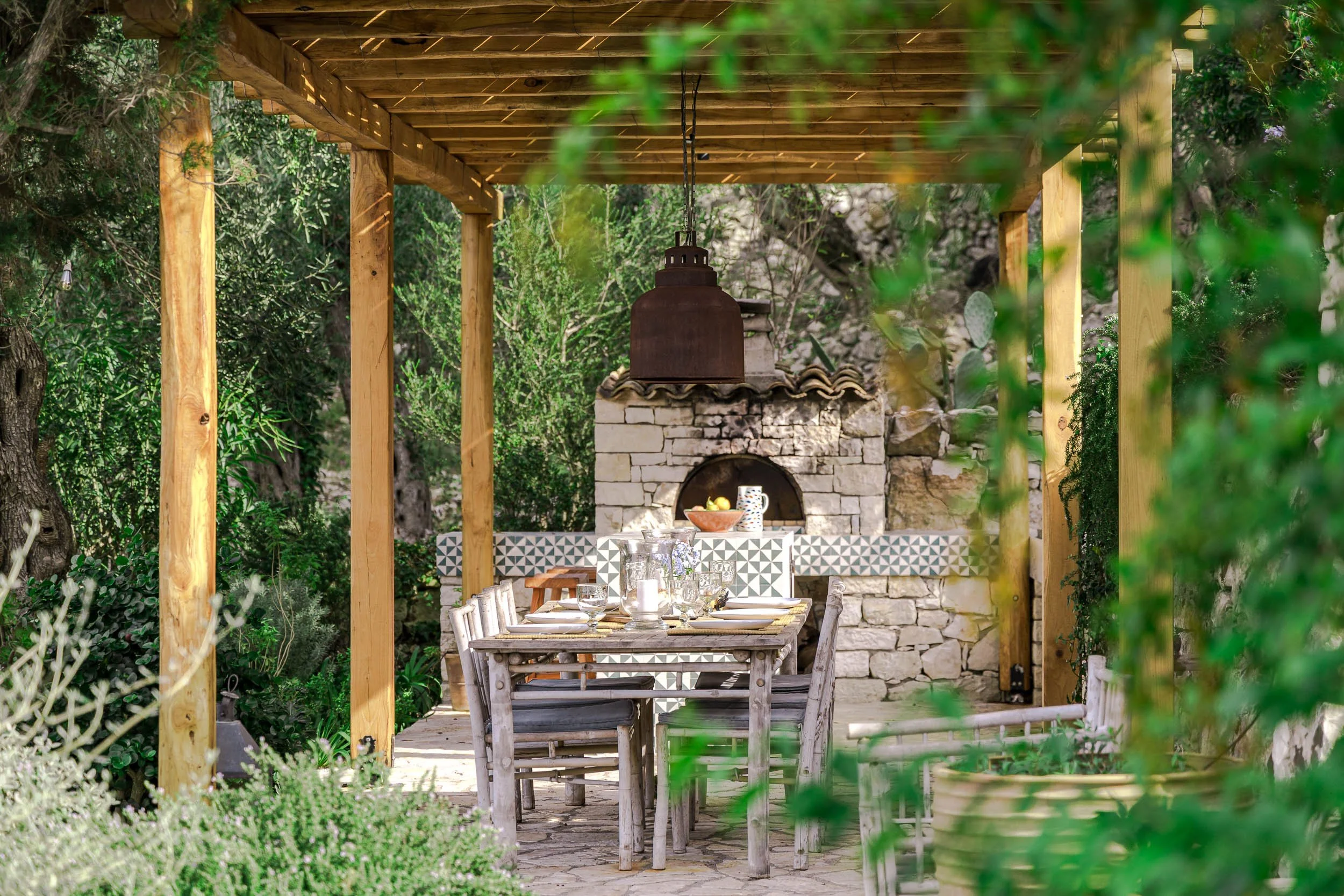 Outdoor patio with a rustic wooden dining table set with glassware, plates, and a centerpiece, under a wooden pergola, with a stone pizza oven in the background, surrounded by lush greenery.