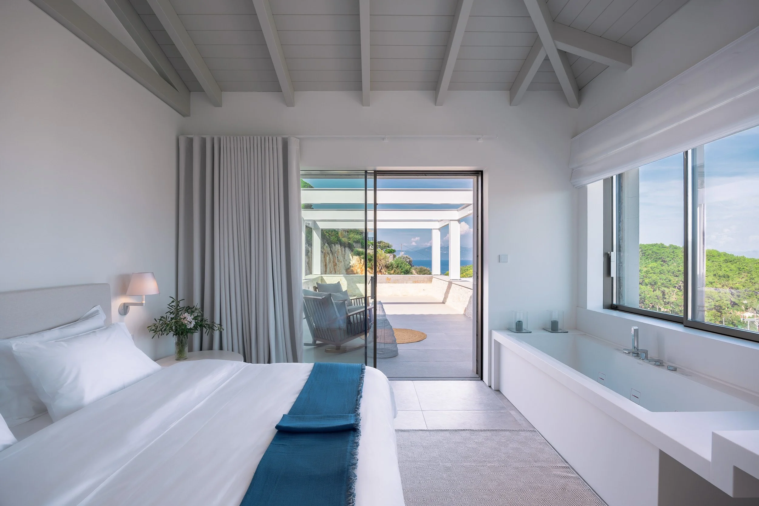A bright bedroom with a large window overlooking green hills, a white bed with a blue throw blanket, a bedside lamp, and a sliding glass door leading to a balcony with outdoor seating and a view of the ocean.