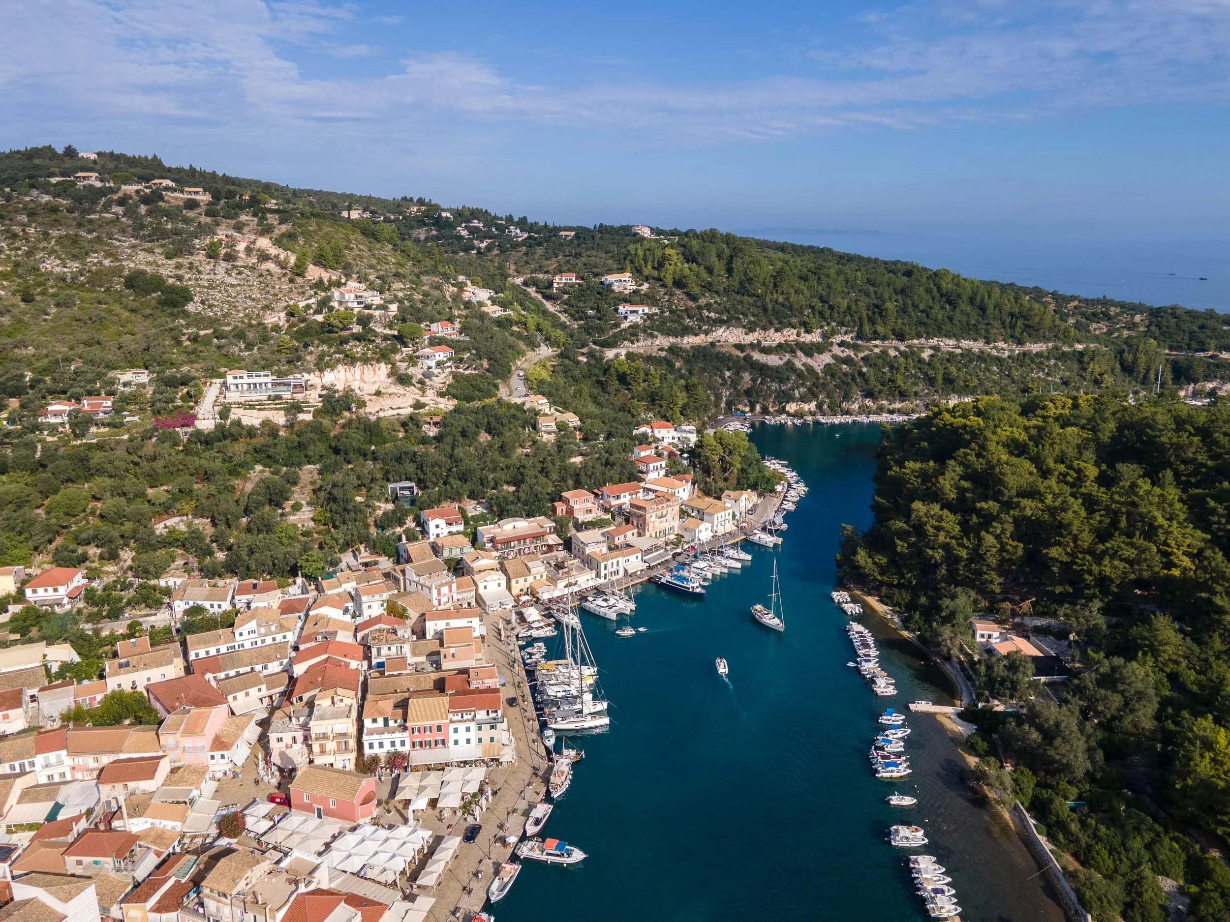 Aerial view of a coastal town with colorful buildings, a marina filled with sailboats, green hills, and a blue sky.