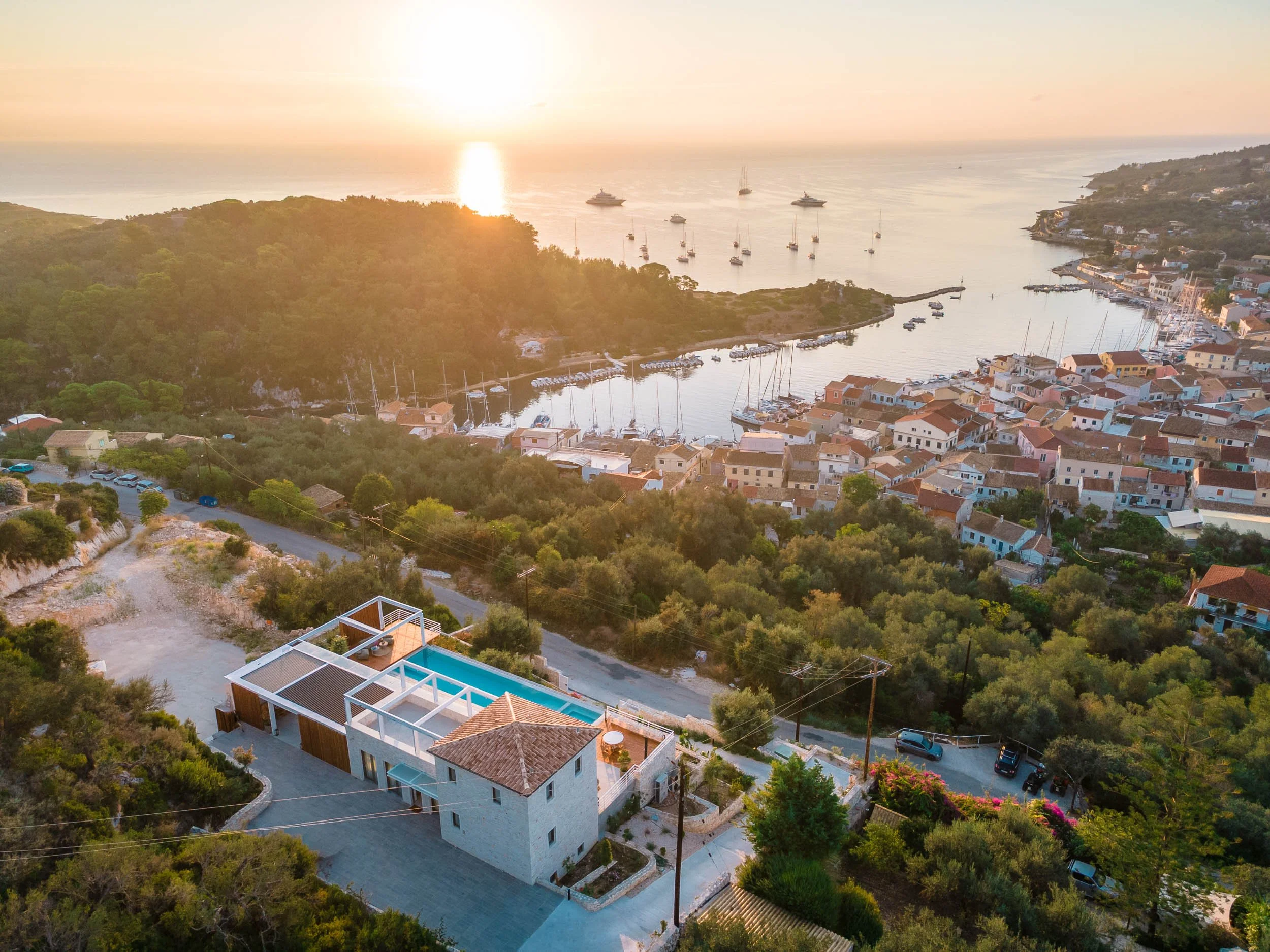 Aerial view of a coastal town at sunset with a pool in the foreground, densely packed houses, green trees, and boats in the harbor.