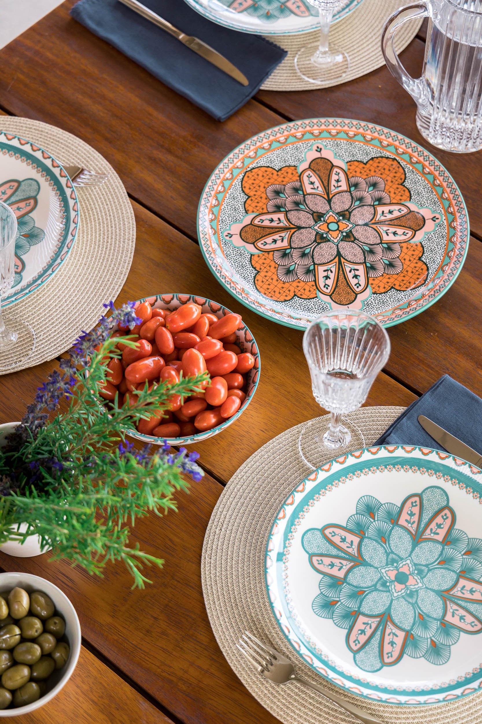 A dining table set with colorful patterned plates, clear glass goblets, beige woven placemats, a navy blue napkin with a knife, and bowls of green and red jelly beans, with a small sprig of purple flowers and greenery in the center.